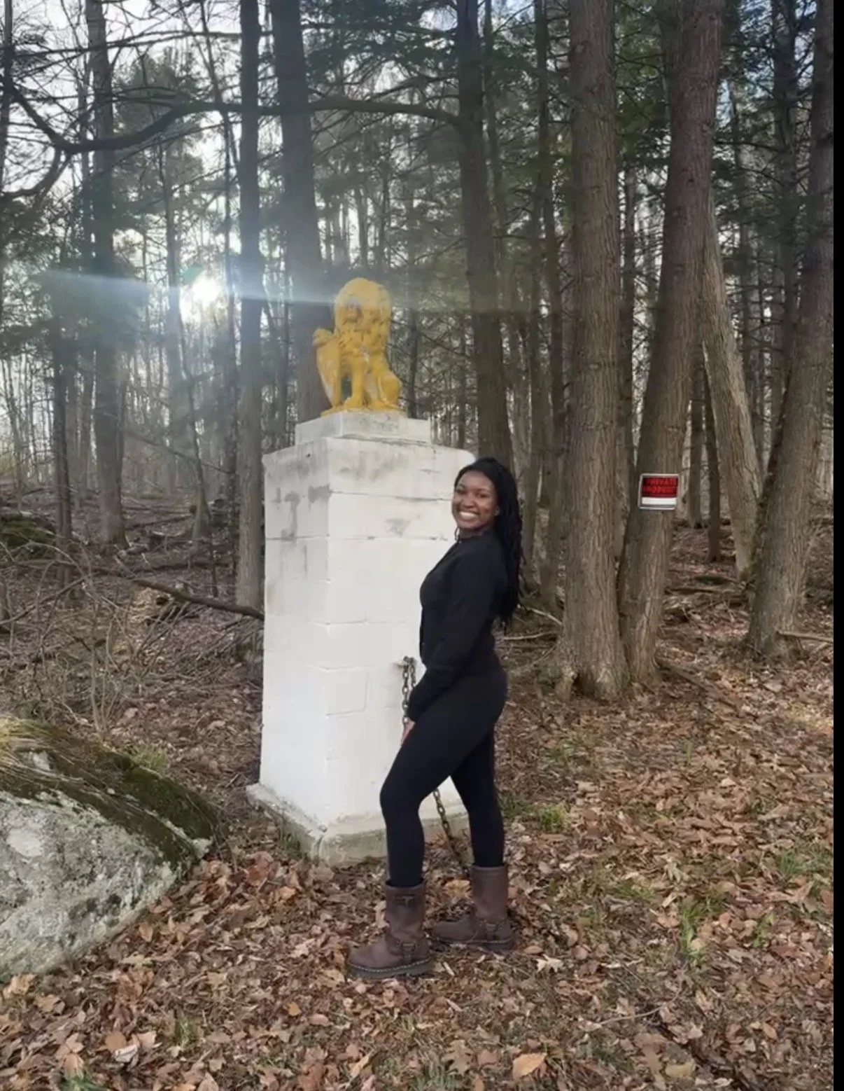 A woman smiling and standing in a wooded area next to a white concrete pedestal with a yellow lion statue on top. There is a small red and white sign on a tree in the background.