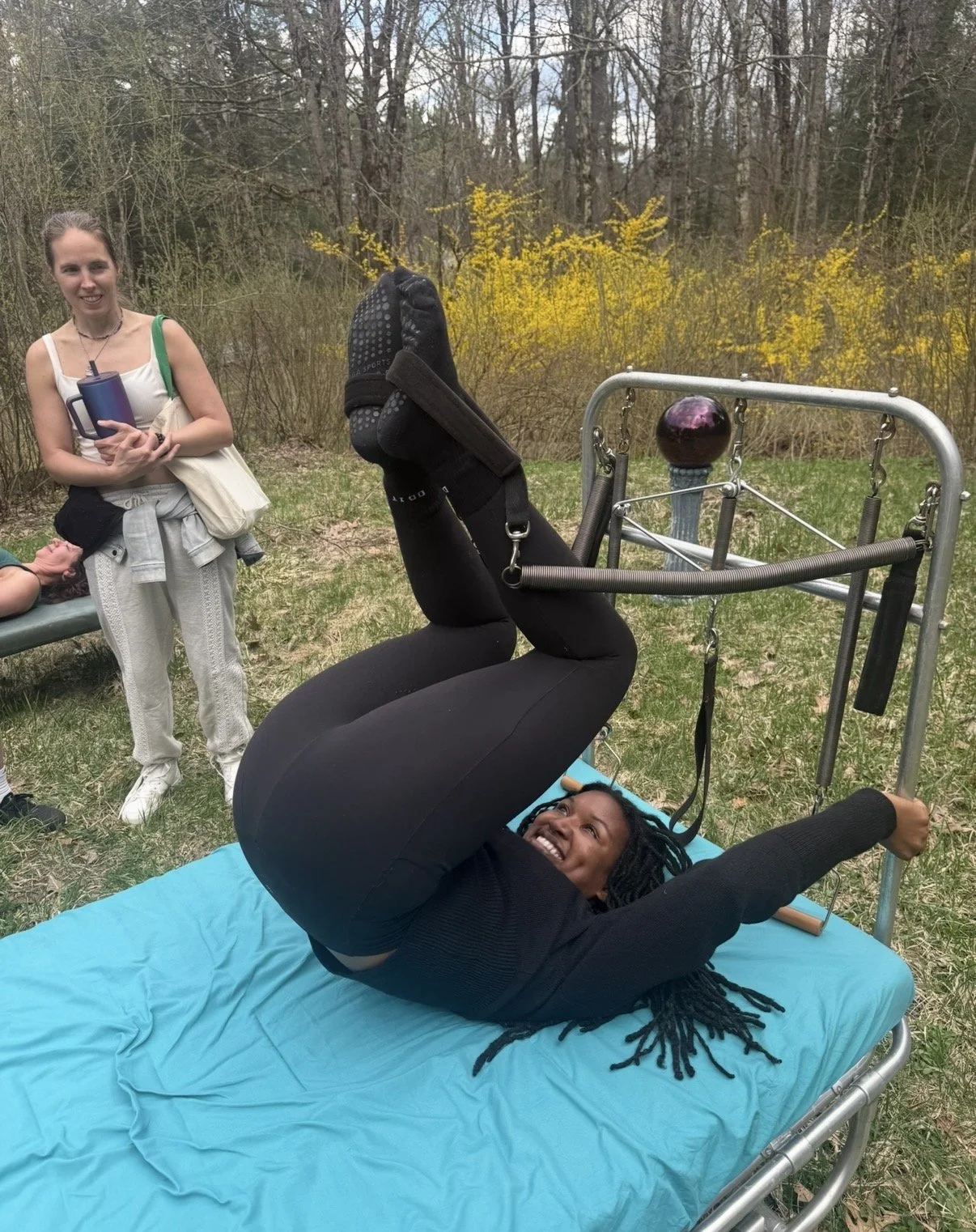 Woman lying on a therapy table outdoors, secured in a harness attached to a metal frame, with people standing nearby watching and smiling.