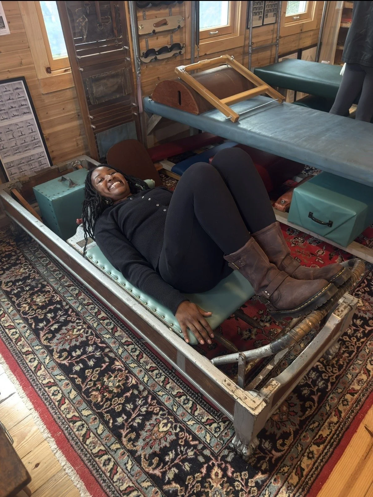 A woman lying on a vintage massage table with a big smile, wearing boots and black clothing, inside a wooden room with exercise or therapy equipment.