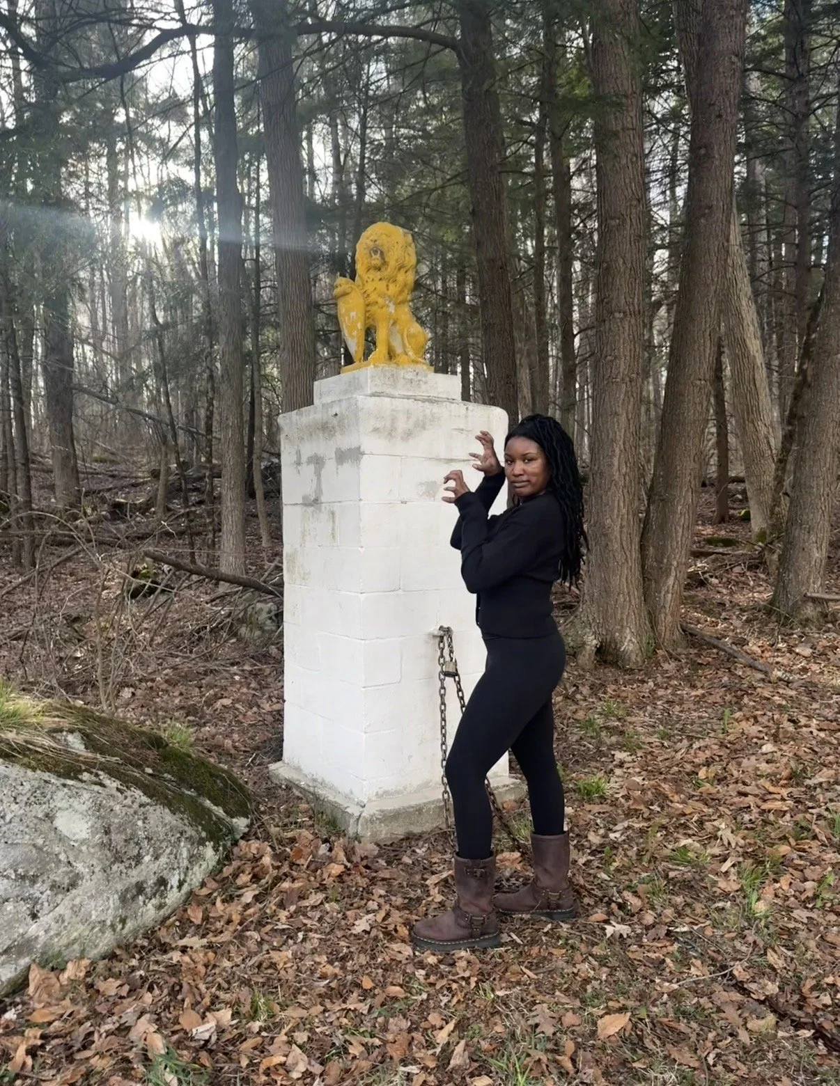 A woman in black clothing and brown boots standing near a white pedestal with a chained lion statue on top, in a wooded area with tall trees and fallen leaves.