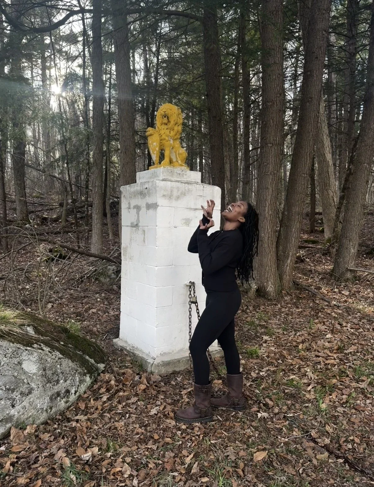 A woman standing in a wooded forest, smiling, dressed in black, posing near a white pedestal with a yellow lion statue on top.