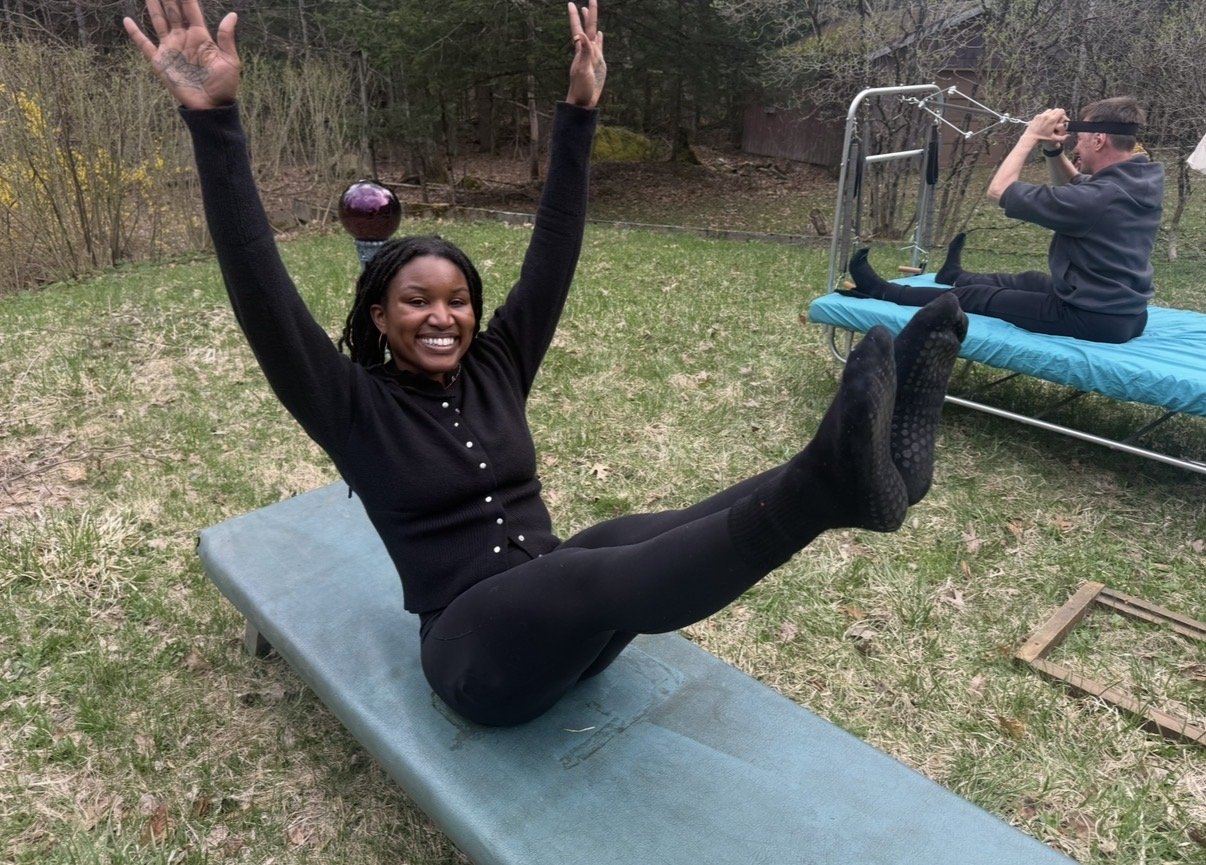 A smiling woman in black clothing sitting on an outdoor exercise mat with arms raised and legs extended, performing a sit-up exercise. In the background, a man is sitting on a similar exercise bed, stretching or looking through a binocular. The scene