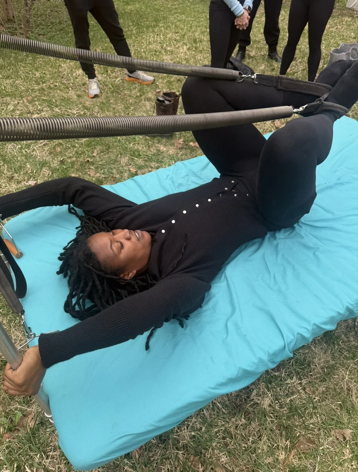 A woman with dreadlocks lying on a blue mat outdoors with her arms stretched above her head while holding onto a metal support frame. Several people are standing nearby, observing the activity.