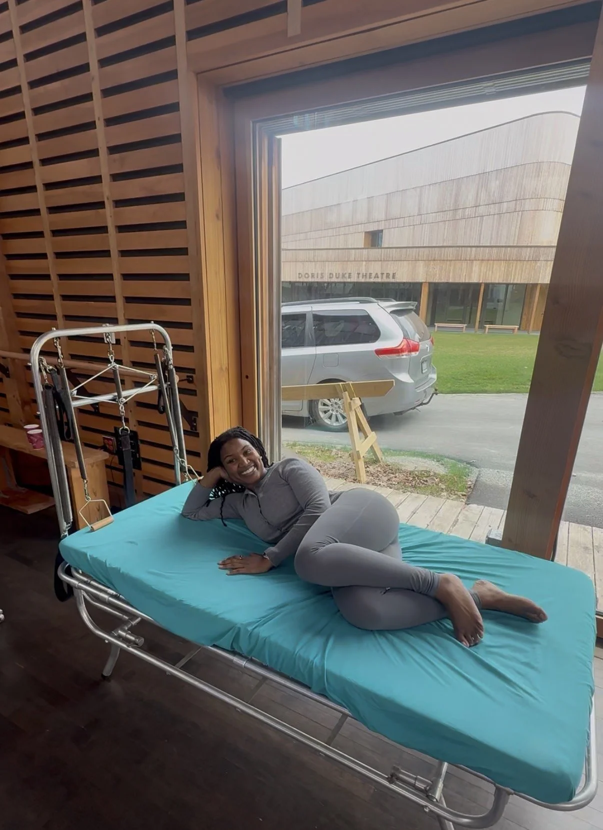 A woman in gray lying on a hospital bed with bright blue sheets, inside a wooden-paneled room. Outside the large open door, there's a parking lot with a gray minivan and a wooden sign.