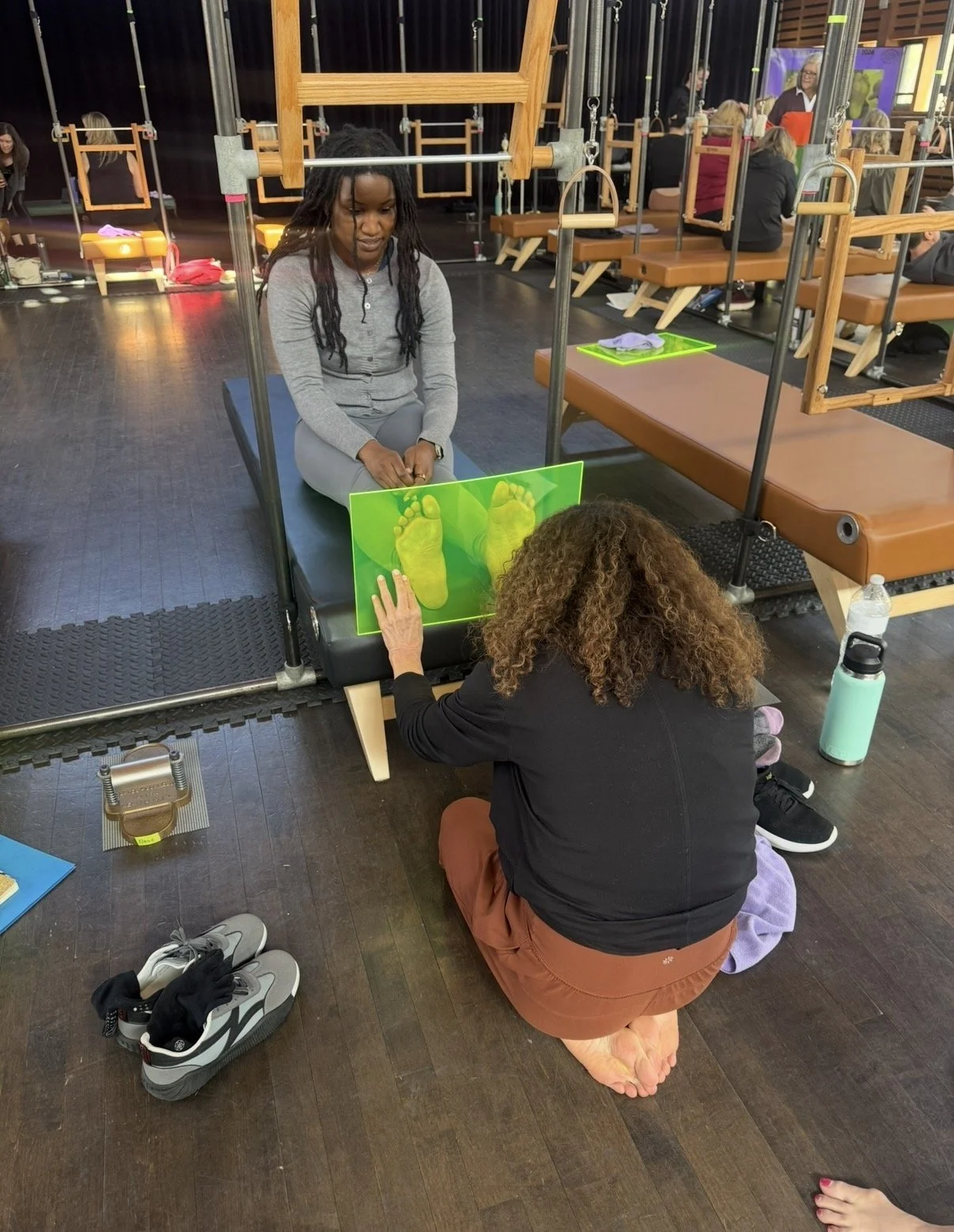 A woman sitting on a black treatment table with her legs straight in front of her, while a therapy professional kneels on the floor holding a green board with yellow footprints. The setting appears to be a therapy or rehabilitation center with multip