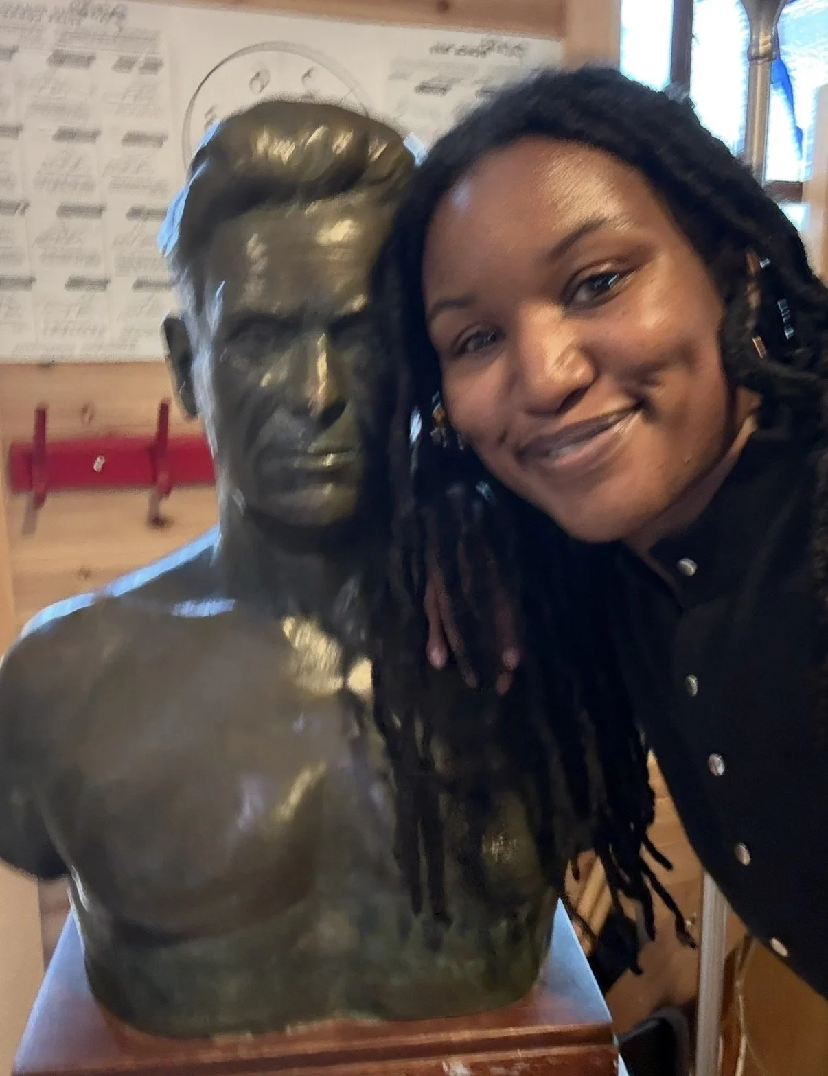 A woman with dark curly hair smiling while leaning close to a bronze bust sculpture of a man with dreadlocks.