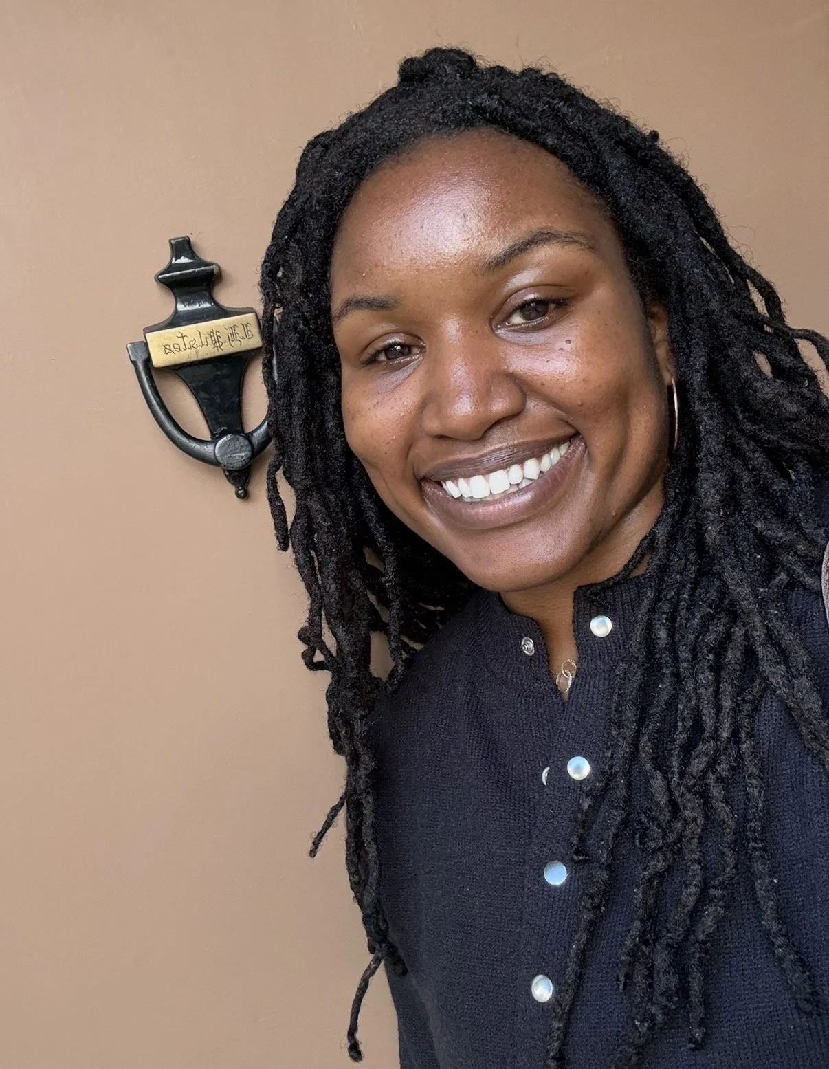 A smiling woman with long dreadlocks and hoop earrings standing in front of a beige wall with a decorative anchor and a gold plaque.