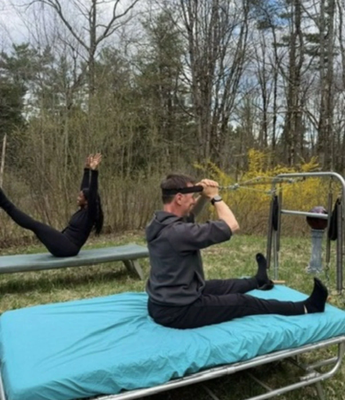 A man sitting cross-legged on an outdoor bed in a backyard, pulling on a resistance band while a woman lying on her back on a bench nearby raises her legs.