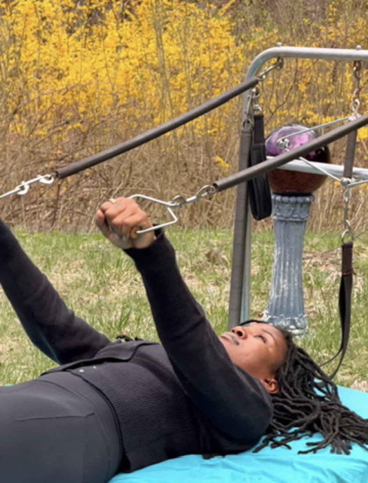 Person lying on the ground outdoors, holding onto a pulley or handle attached to a metal frame, with yellow and orange autumn trees in the background.
