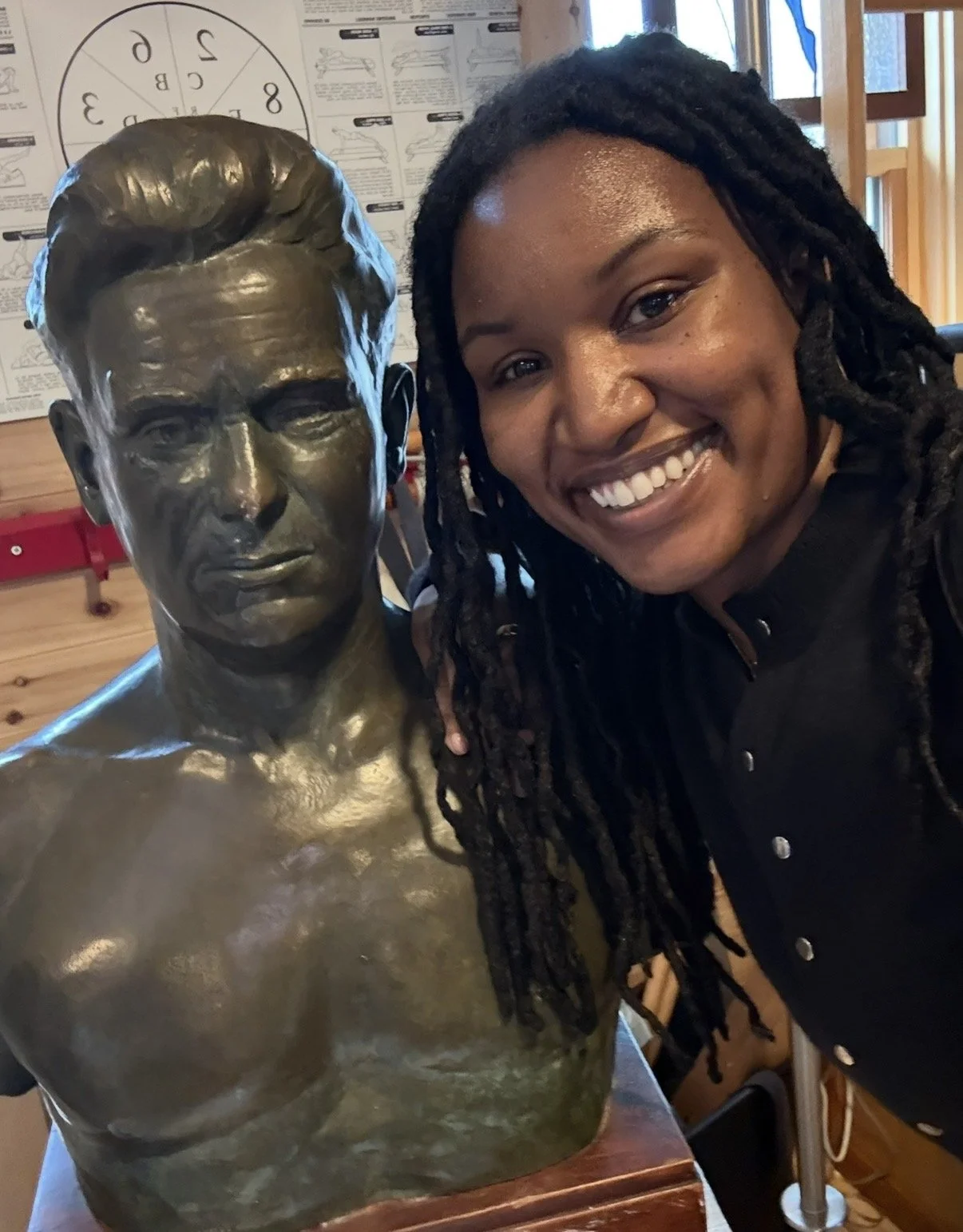 A woman with long dreadlocks smiling next to a bronze bust sculpture of a person's head and shoulders in a wooden-paneled room.