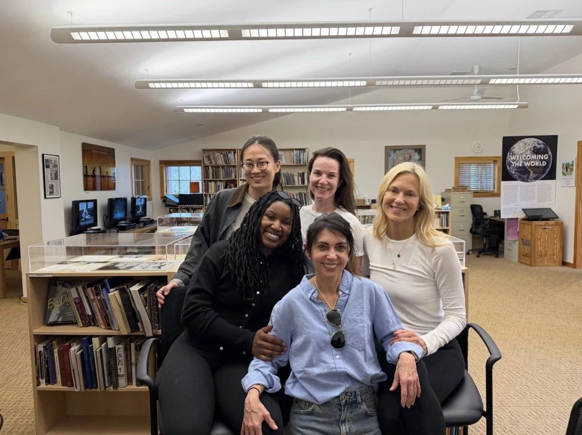 A group of five women posing for a photo inside a library or community center, with bookshelves, computers, and posters visible in the background.