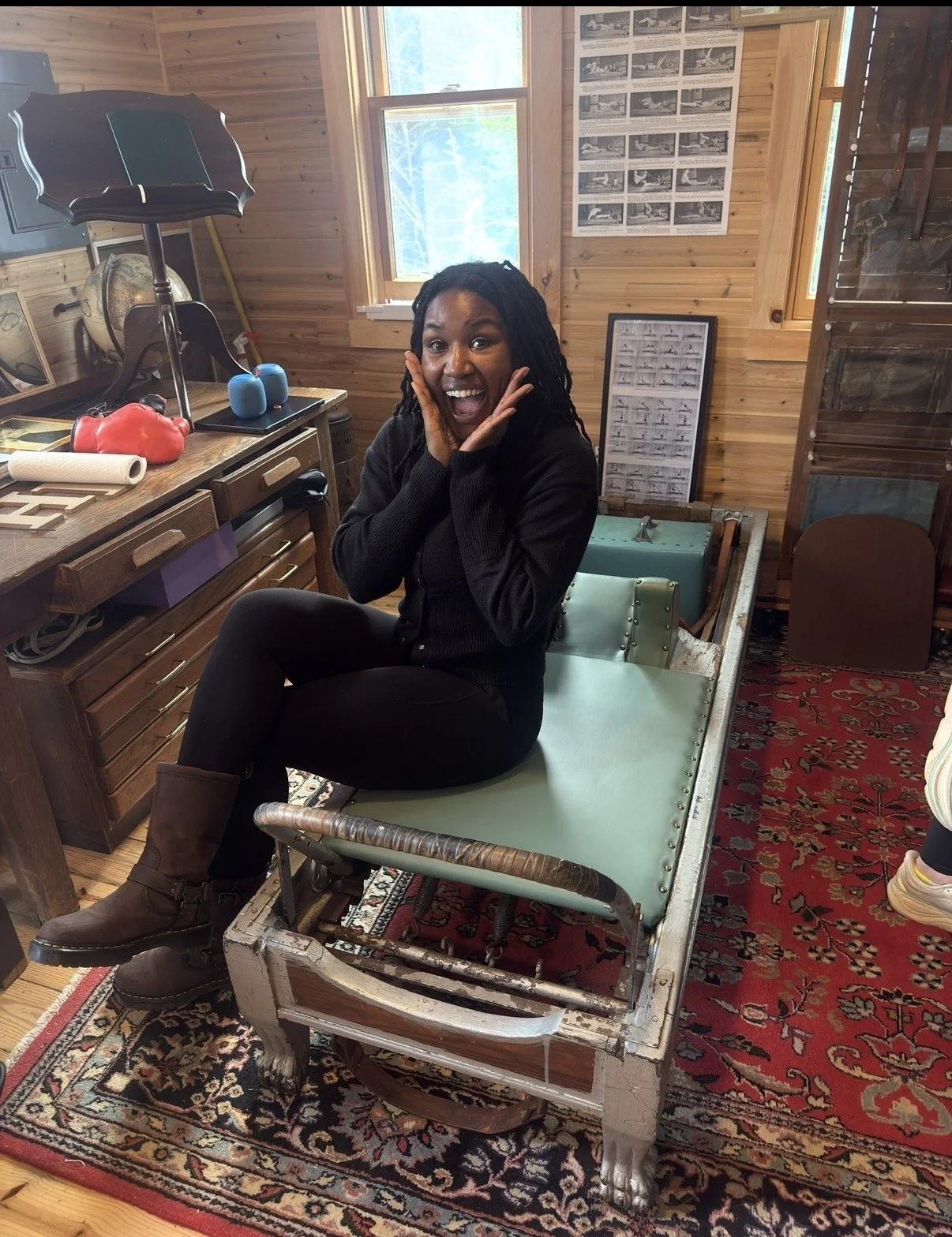 A woman sitting on an antique hospital bed in a wooden-paneled room, smiling with hands on her face, surrounded by vintage decor including a lamp, framed pictures, and a patterned rug.