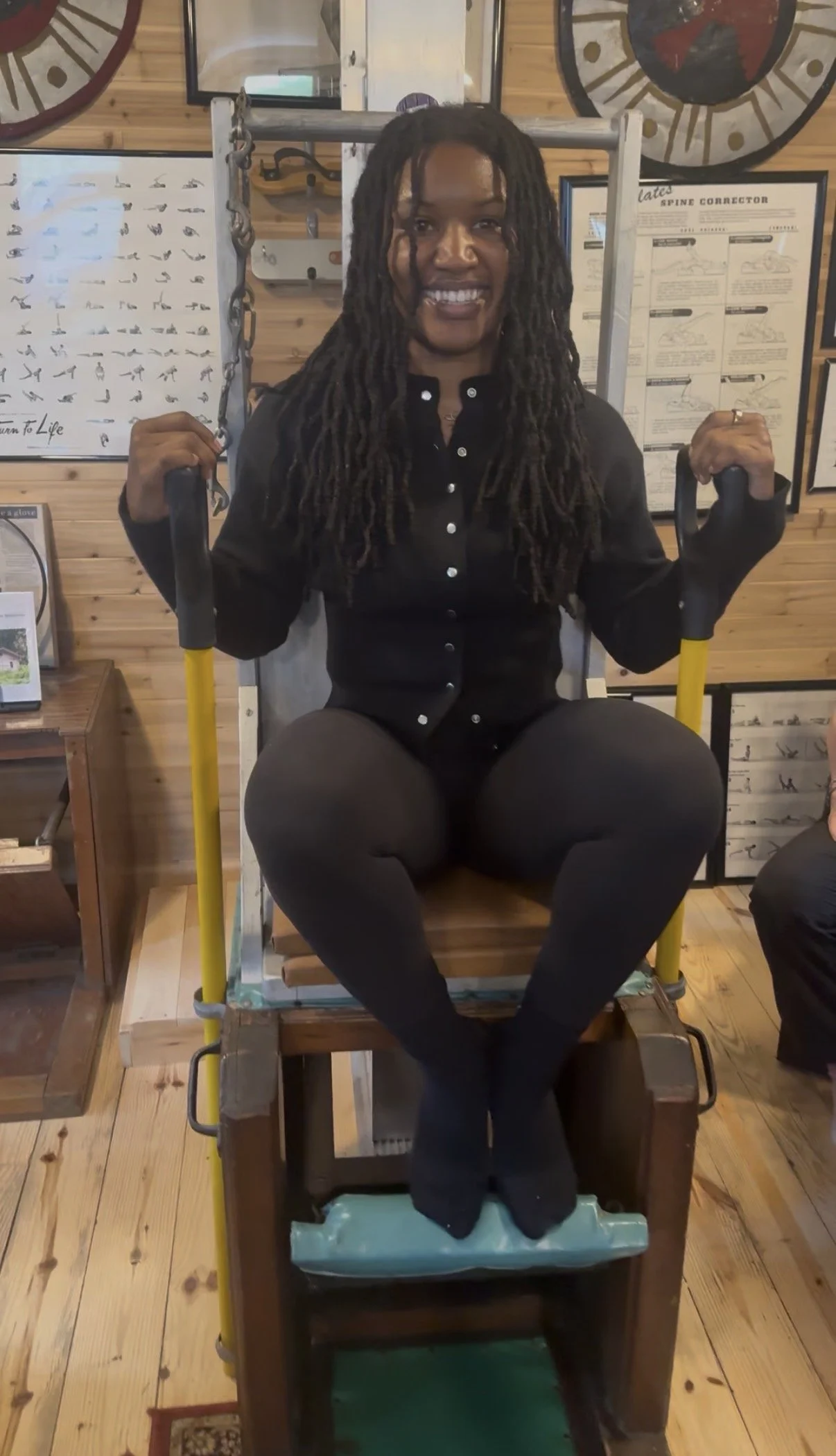 A woman with dreadlocks sitting on a wooden chair with her feet in a foot soaking tank, holding onto a metal support bar in a spa or wellness center, with massage charts on the wall behind her.