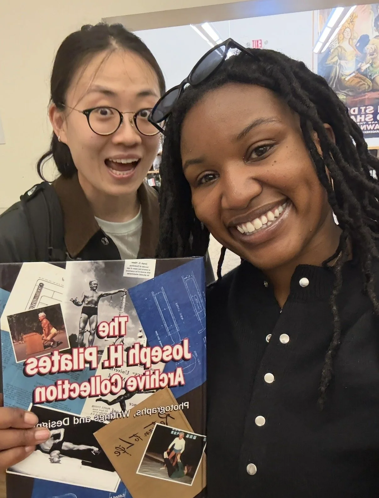 Two women smiling and taking a selfie inside a building, one holding a brochure for Joseph H. Pulaski Academy and College. The woman on the right has glasses on her head and the woman on the left is wearing glasses and has short dark hair.