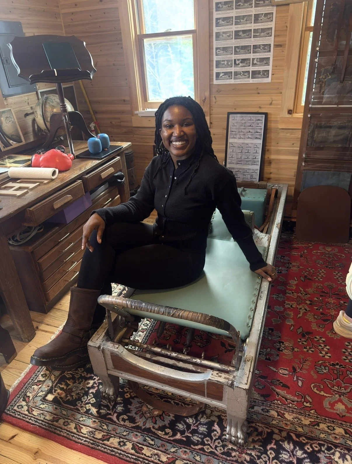 A woman with dreadlocks, smiling, sitting on a vintage medical examination table in a wooden-paneled room.
