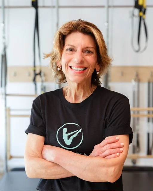 A smiling woman trainer at Providence pilates center in a black T-shirt with 'Providence Pilates' logo, standing with hands on hips in a studio setting.