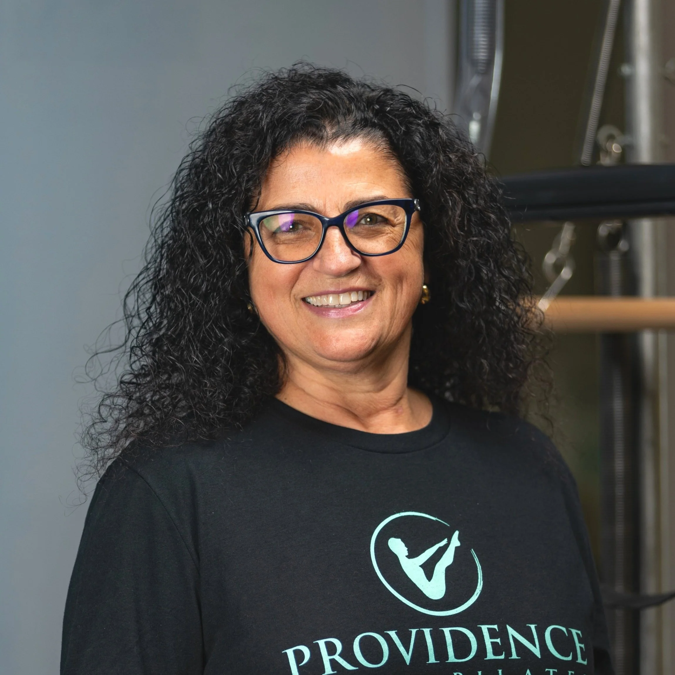 Woman trainer at Providence pilates center with curly black hair and glasses smiling, wearing a black Providence t-shirt, in an indoor setting with gym equipment in the background.