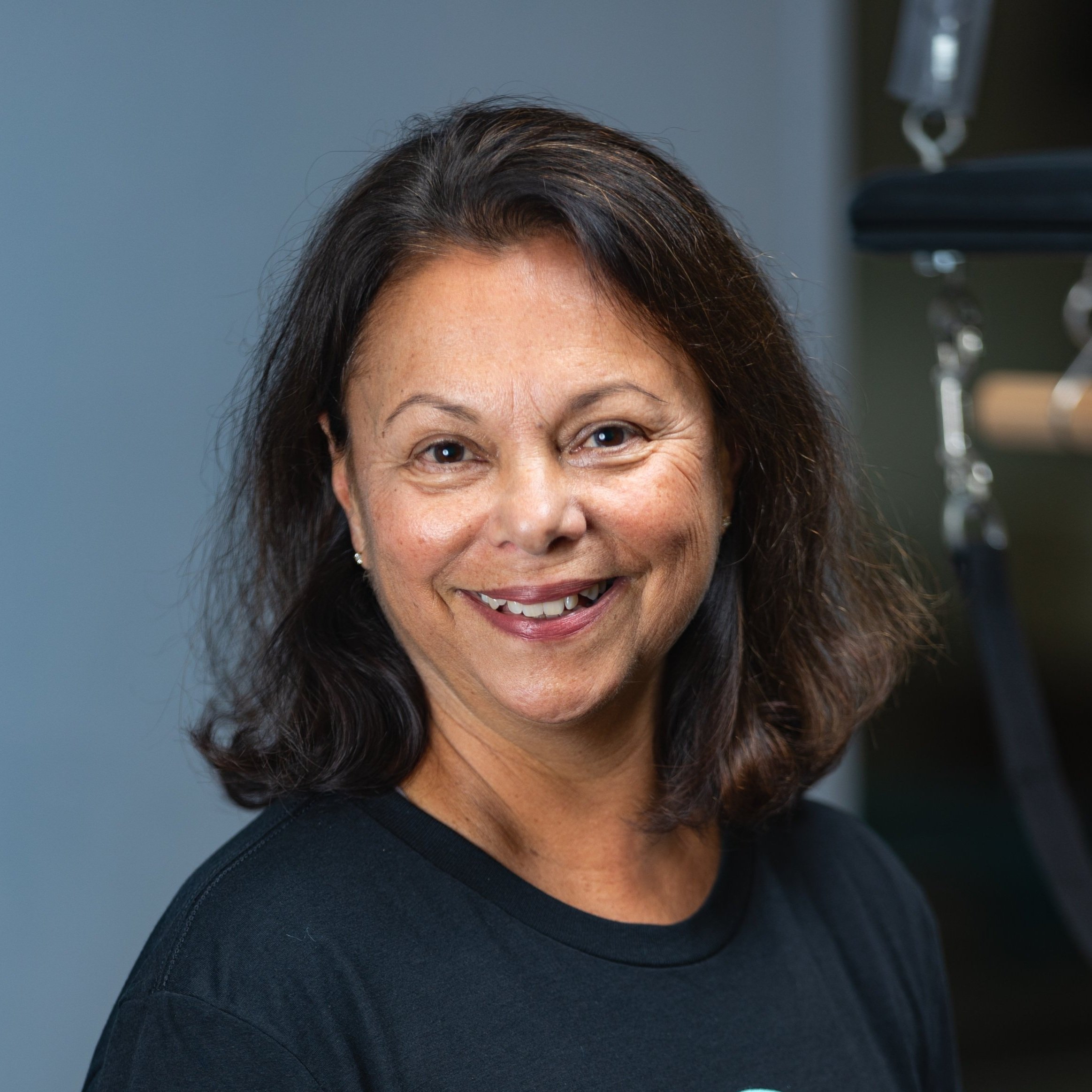 Smiling middle-aged woman  trainer at Providence pilates center with dark hair in a black shirt standing in a room with exercise equipment.