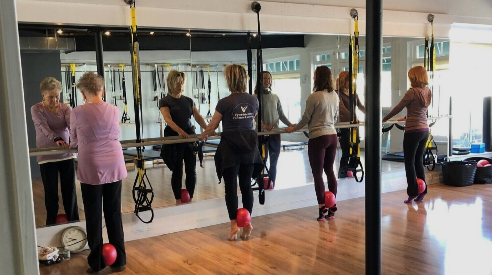 Group of women in a pilates fitness class practicing barre exercises in a gym with suspension straps, mirrors, and wooden floors.