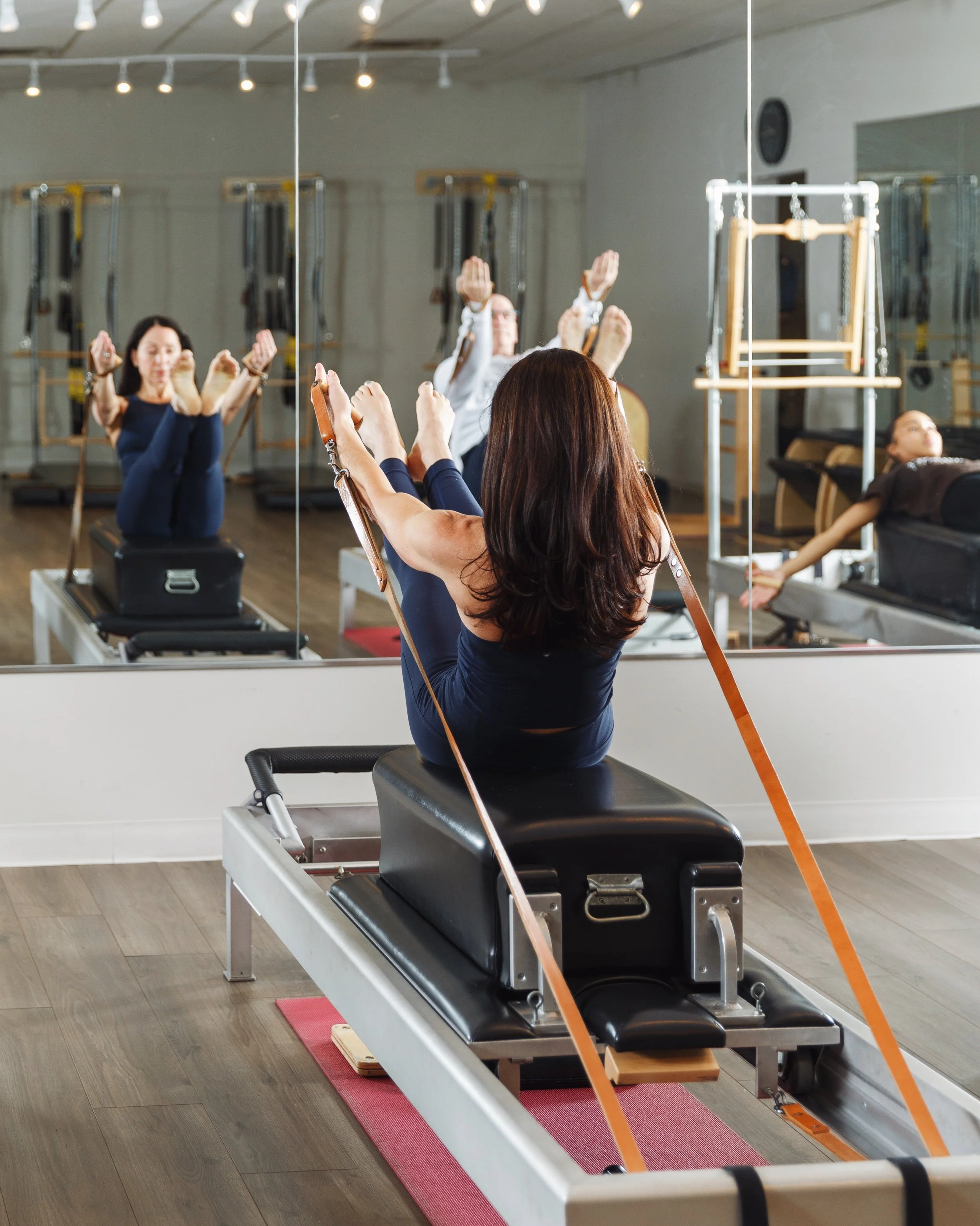 A woman practices classic Pilates on a reformer machine in a mirrored studio, stretching her legs while holding straps.