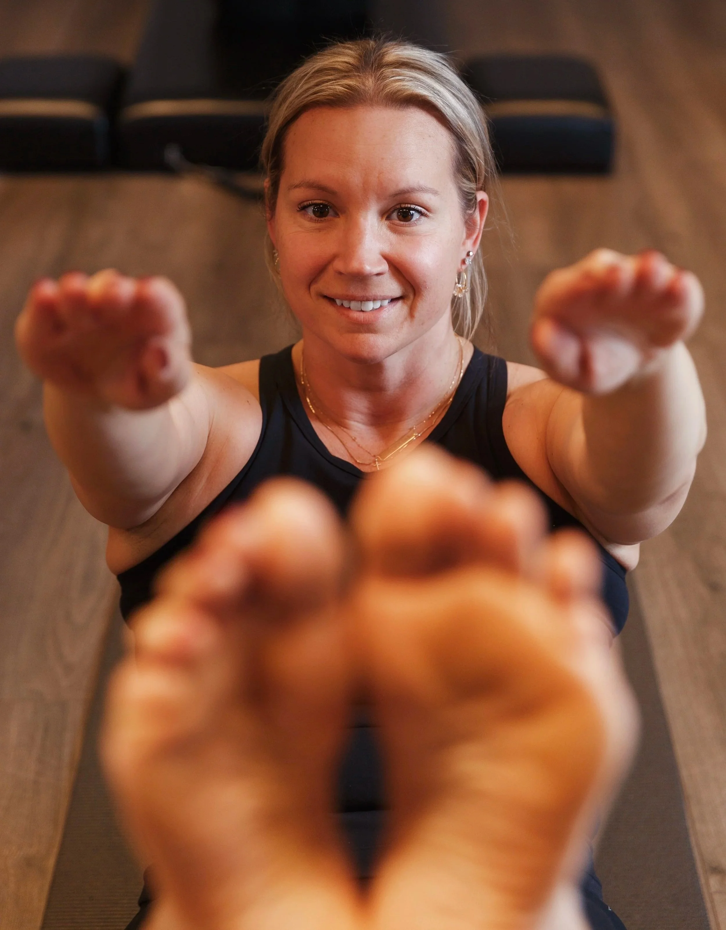 Woman in power pilates workout clothes sitting on a mat, arms extended forward, with her feet in the foreground, smiling at the camera.