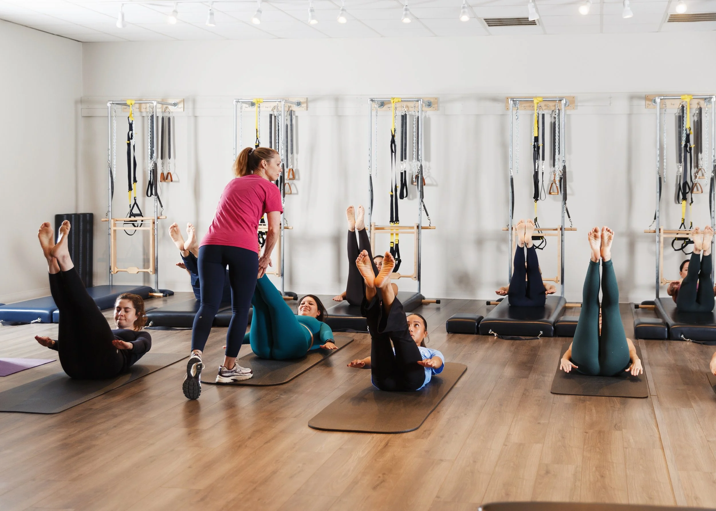 Group of women participating in a Pilates class, lying on mats with legs raised, guided by an instructor in a fitness studio with Pilates equipment in the background.