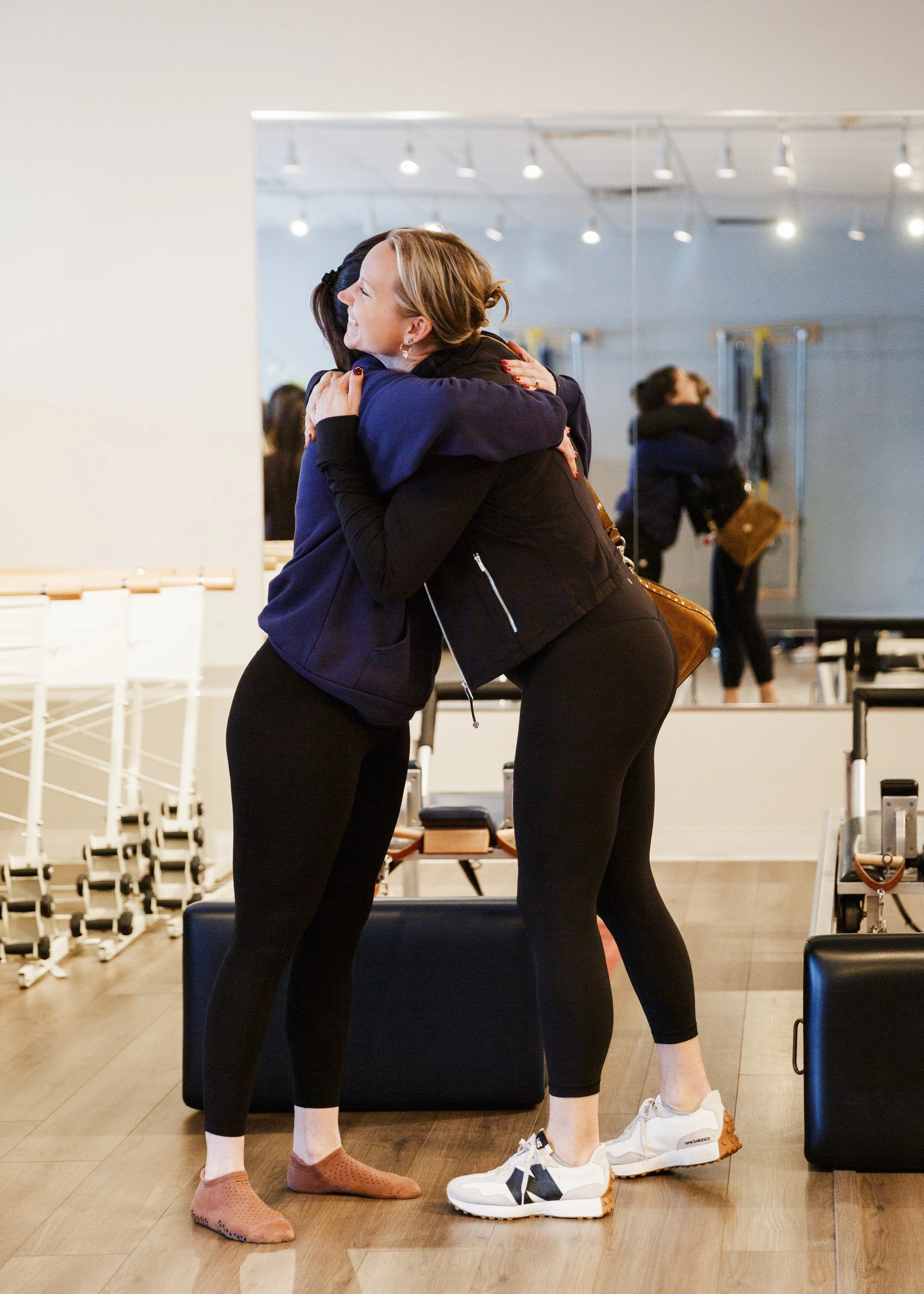 Two women who practice pilates fitness hugging in a gym, wearing workout clothes, in front of a large mirror.