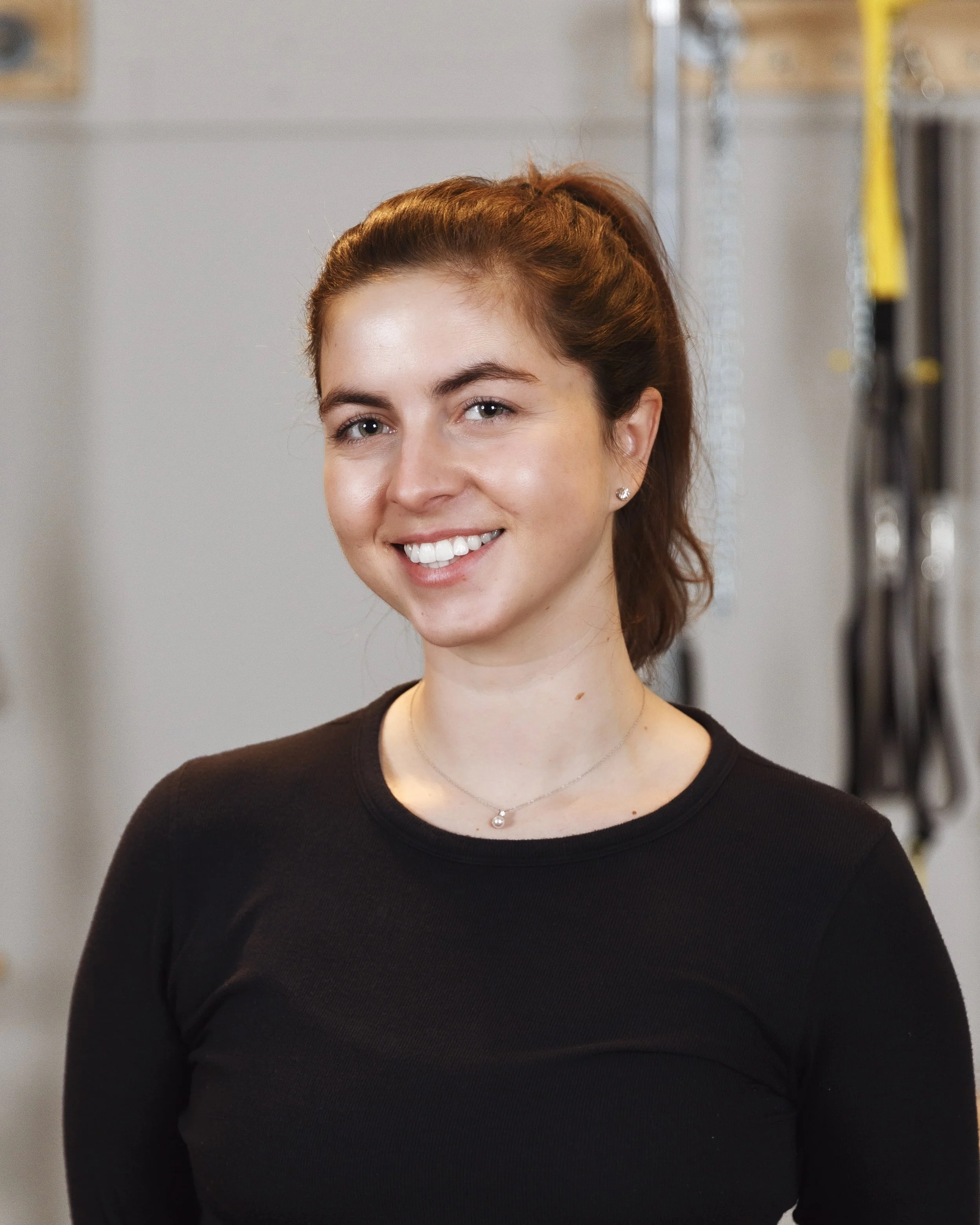 A young woman trainer at Providence pilates center with brown hair tied back, smiling, wearing a black top and a silver necklace, standing in an indoor setting with gray walls and equipment in the background.