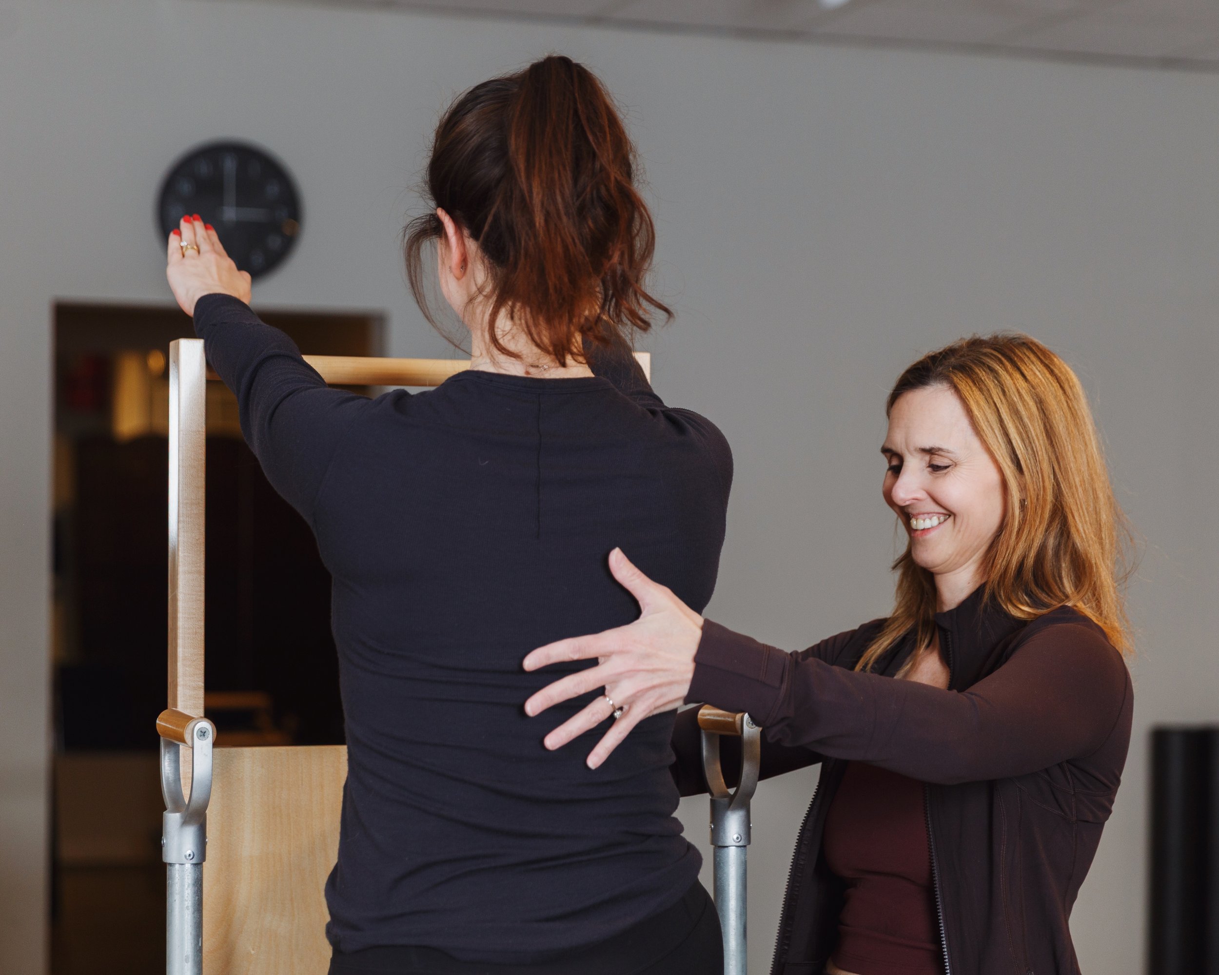 A power pilates trainer woman assists another woman with an exercise on pilates fitness equipment in a bright room; both are wearing black clothing.