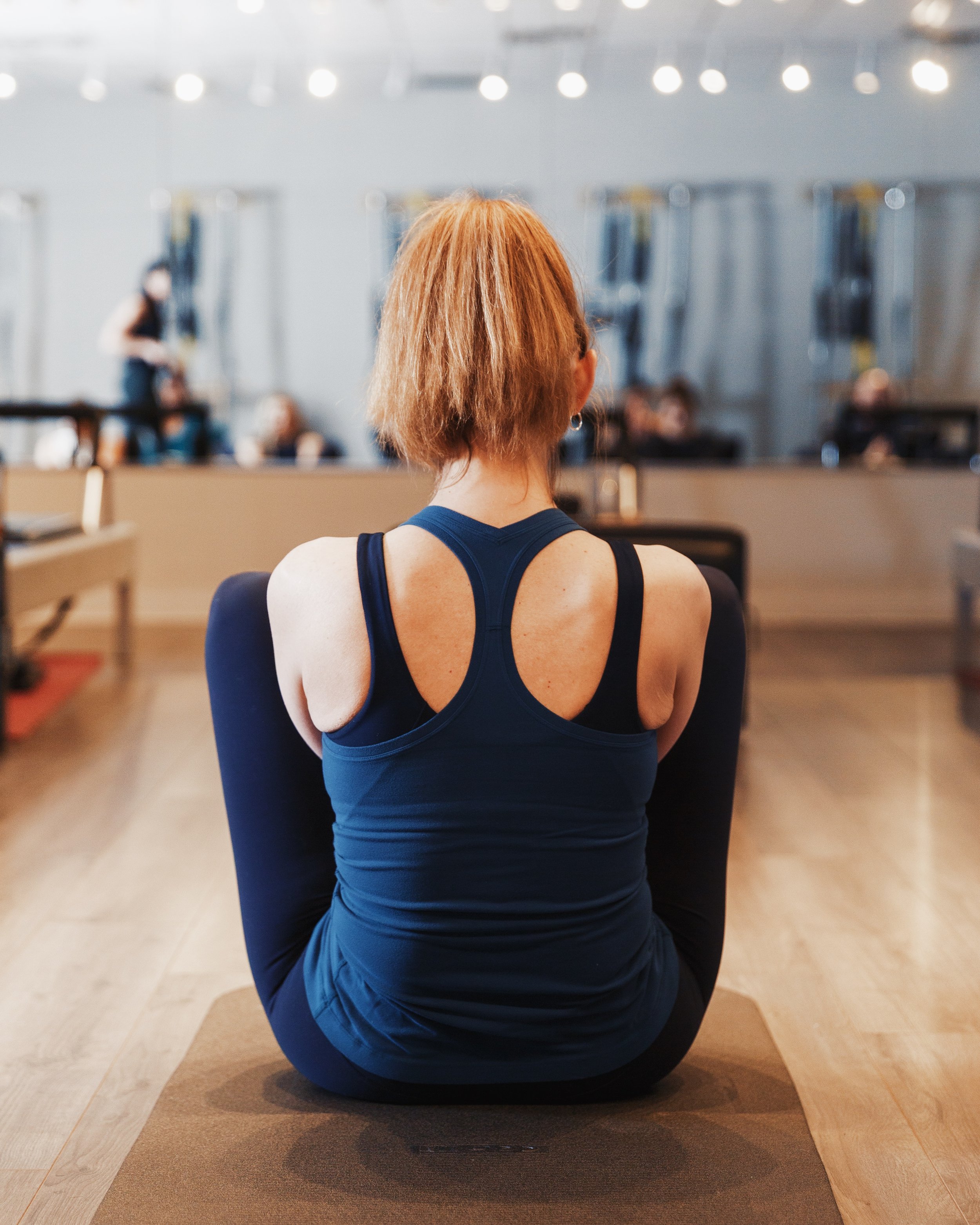 Back view of a woman with short red hair seated on a yoga mat in a cumberland pilates fitness studio, wearing a black sports outfit, with other people and exercise equipment visible in the background.