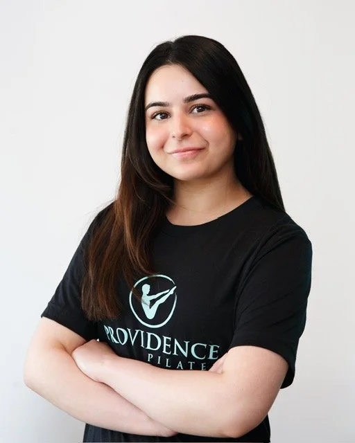 A young woman trainer at Providence pilates center with dark hair and a black Providence t-shirt, standing with arms crossed and smiling in an indoor gym or studio.