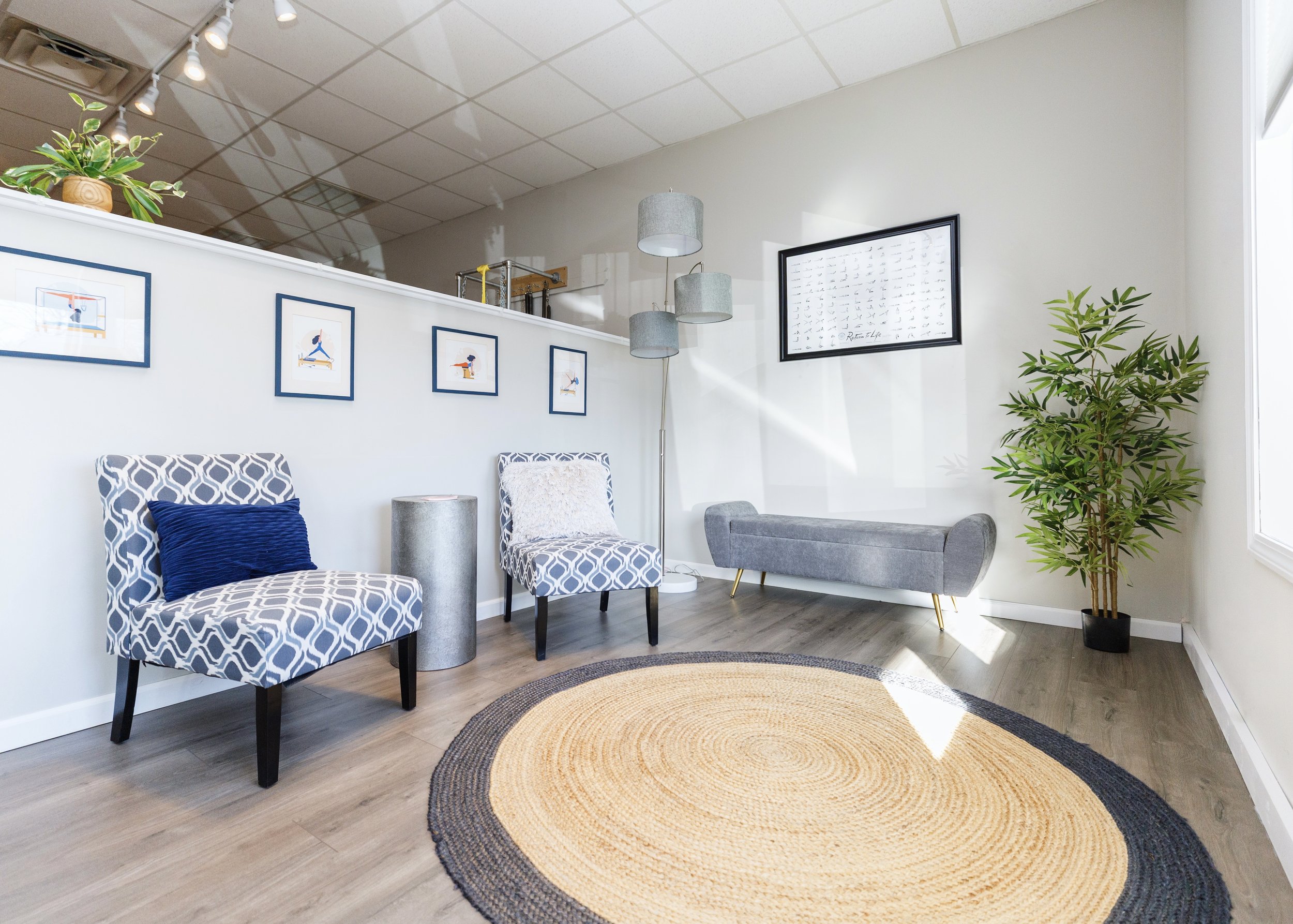 A bright waiting room at Providence pilates center with patterned chairs, a gray bench, a tall floor lamp, framed artwork, a potted plant, and a round woven rug on wood flooring.