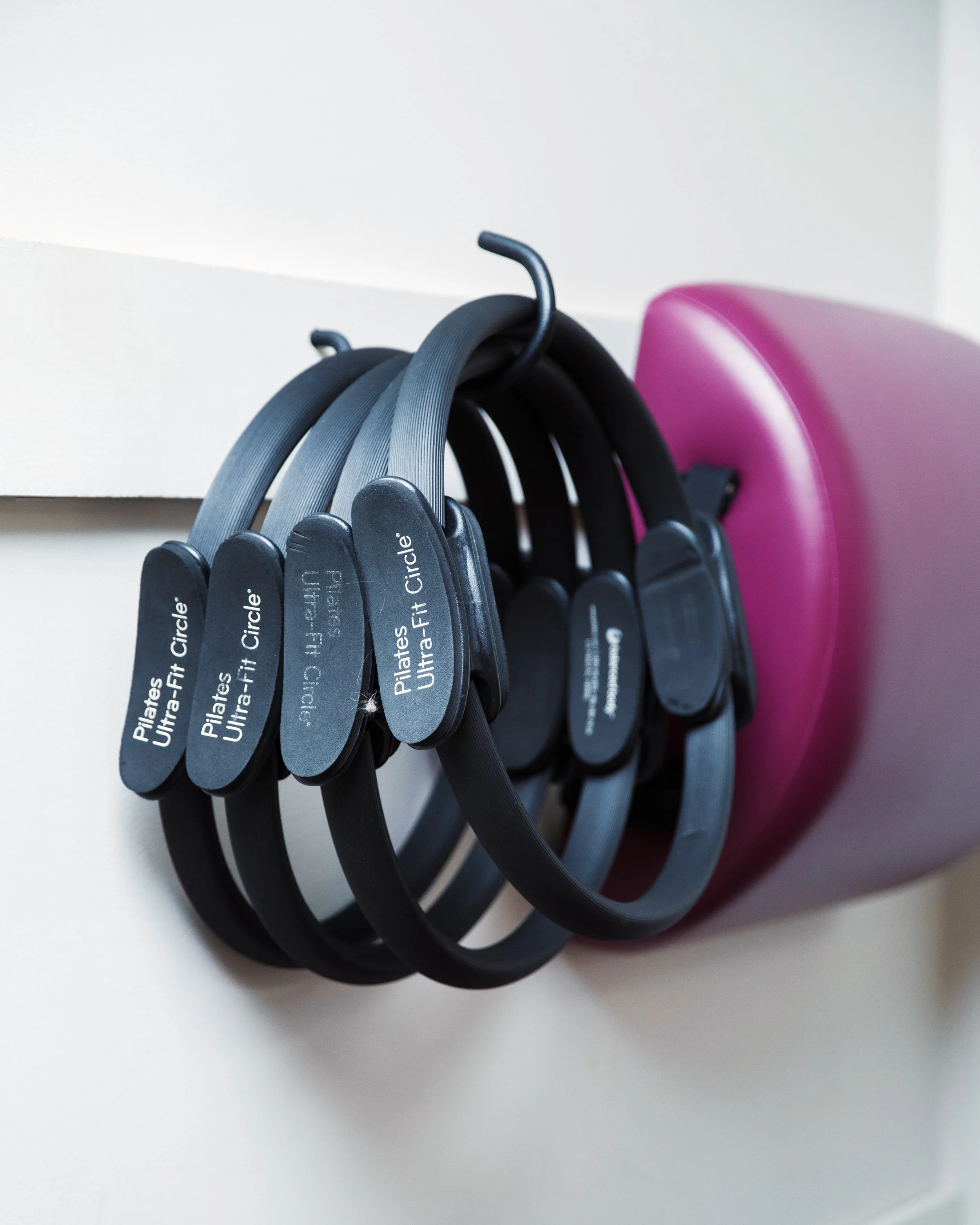 A set of black and gray Pilates rings hanging on hooks on a white wall, with a pink exercise roller partially visible behind them.