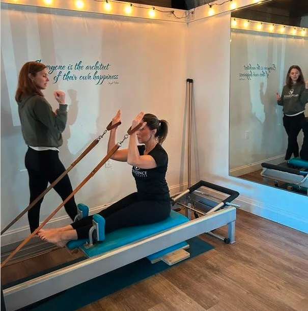 Woman performing Pilates exercises on a reformer machine in a studio, with an instructor standing nearby and a mirror reflecting the scene, warm lighting, and wall quote about happiness.
