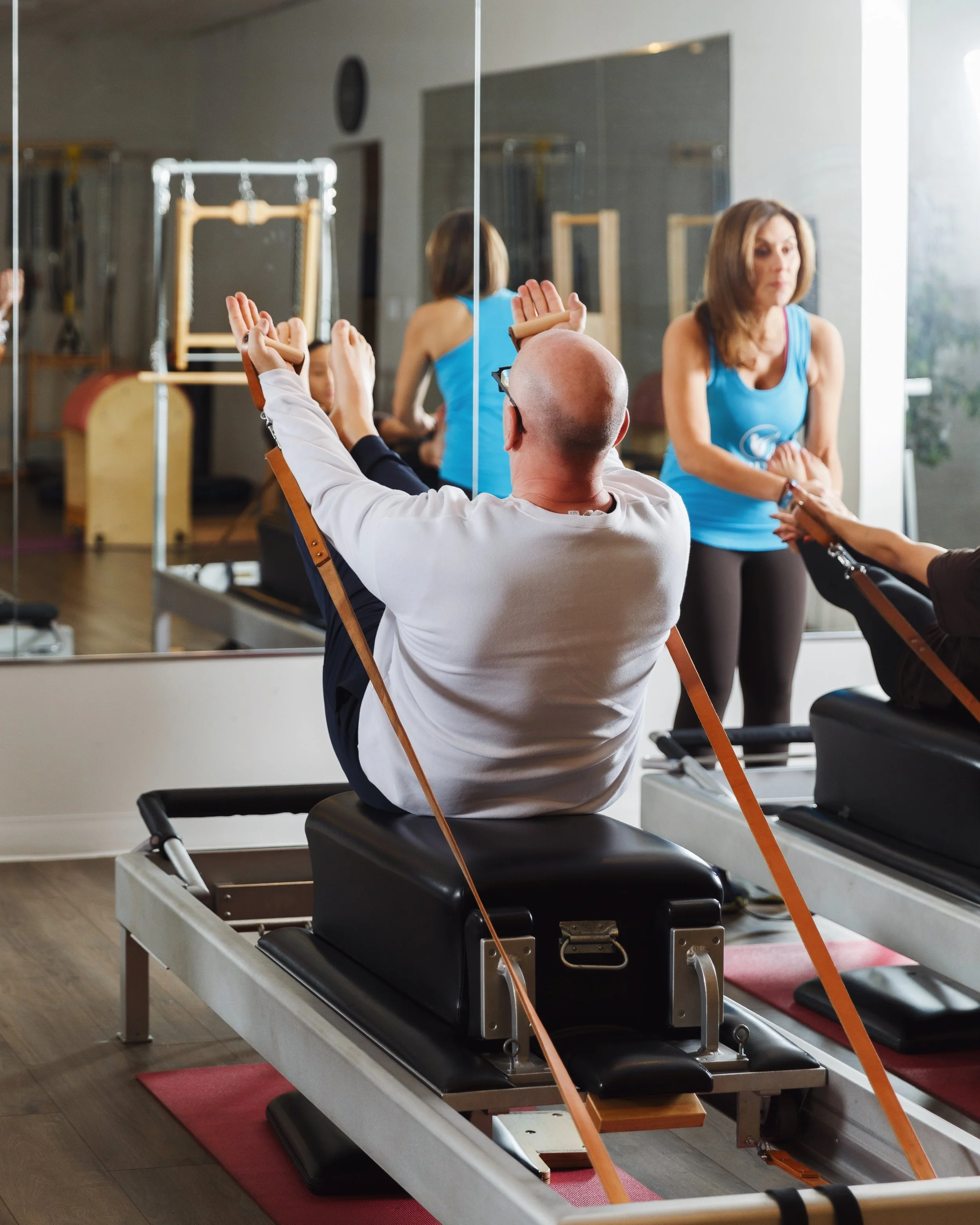 A man doing physical fitness exercise on a Pilates reformer machine while a woman pilates trainer in a blue shirt supervises his class in a providence pilates gym with mirrors.