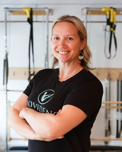 A woman trainer at Providence pilates center smiling, wearing a black Providence Pilates t-shirt, in a fitness studio with workout equipment in the background.
