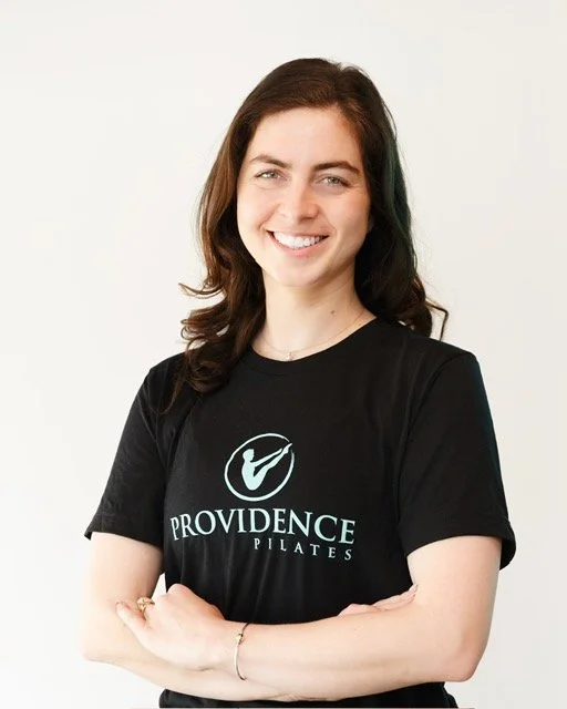 A young woman trainer at Providence pilates center with brown hair tied back, smiling, wearing a black top and a silver necklace, standing in an indoor setting with gray walls and equipment in the background.