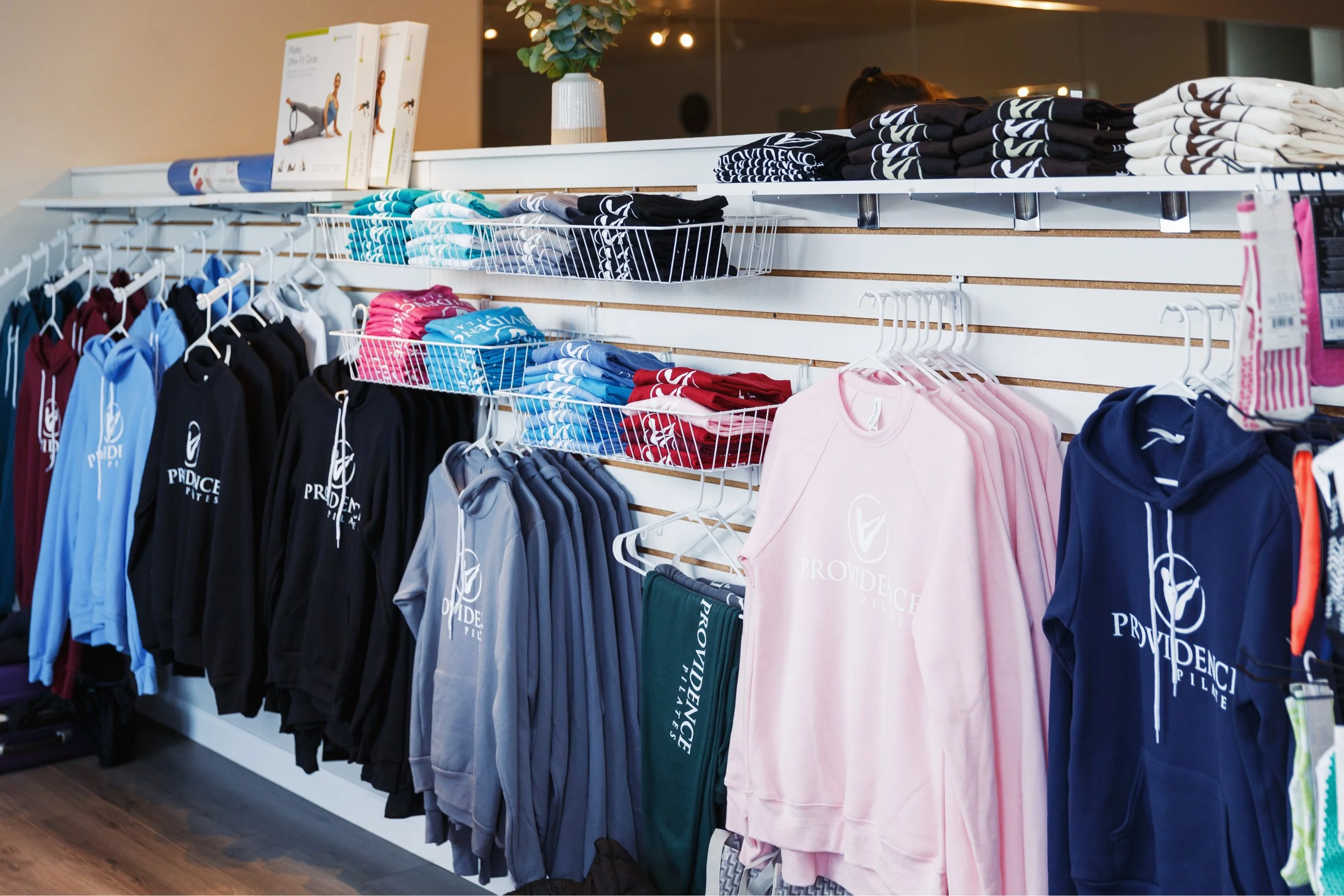 Providence pilates Clothing store display with hoodies and t-shirts, arranged on racks and shelves, with a potted plant on top.