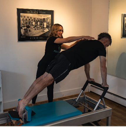 A woman providence pilates trainer assists a man on a Pilates reformer machine in a studio.
