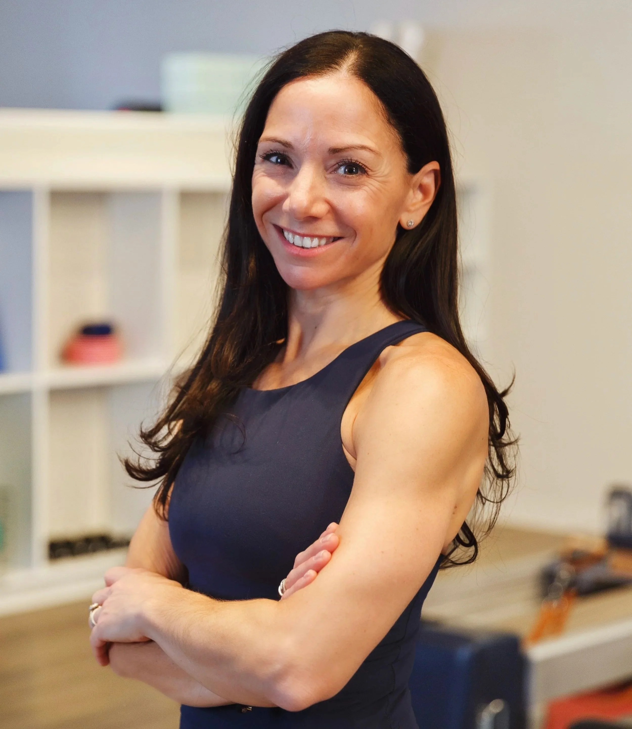 A woman trainer at Providence pilates center with long dark hair smiling and crossing her arms, standing indoors in front of shelving and blurred background.