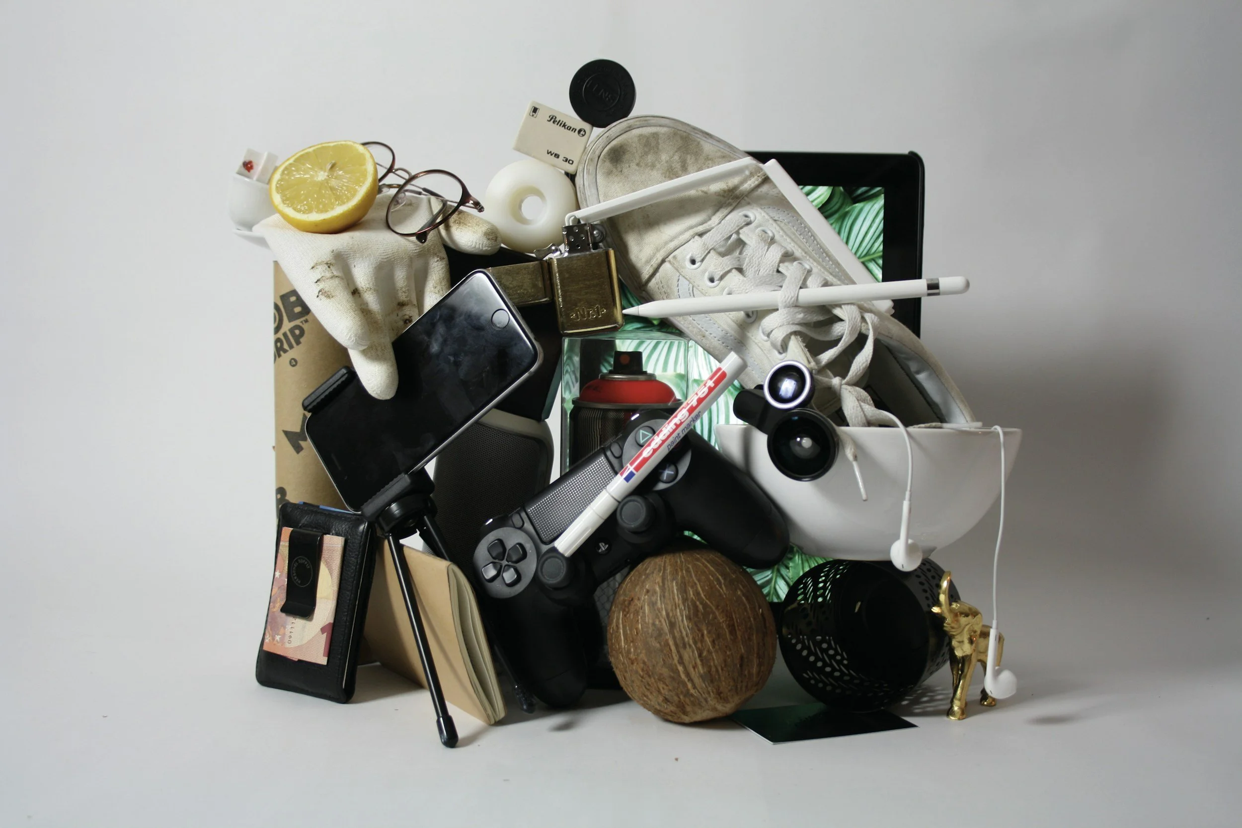 A pile of various objects including a lemon half, glasses, a white glove, a smartphone, a cassette tape, a small speaker, a portable charger, a spray can, a gaming controller, a tennis ball, a coconut, a black decorative item, a white mug, a white ballpoint pen, a tablet, a small gold figurine, earphones, and a white sneaker, arranged on a plain background.
