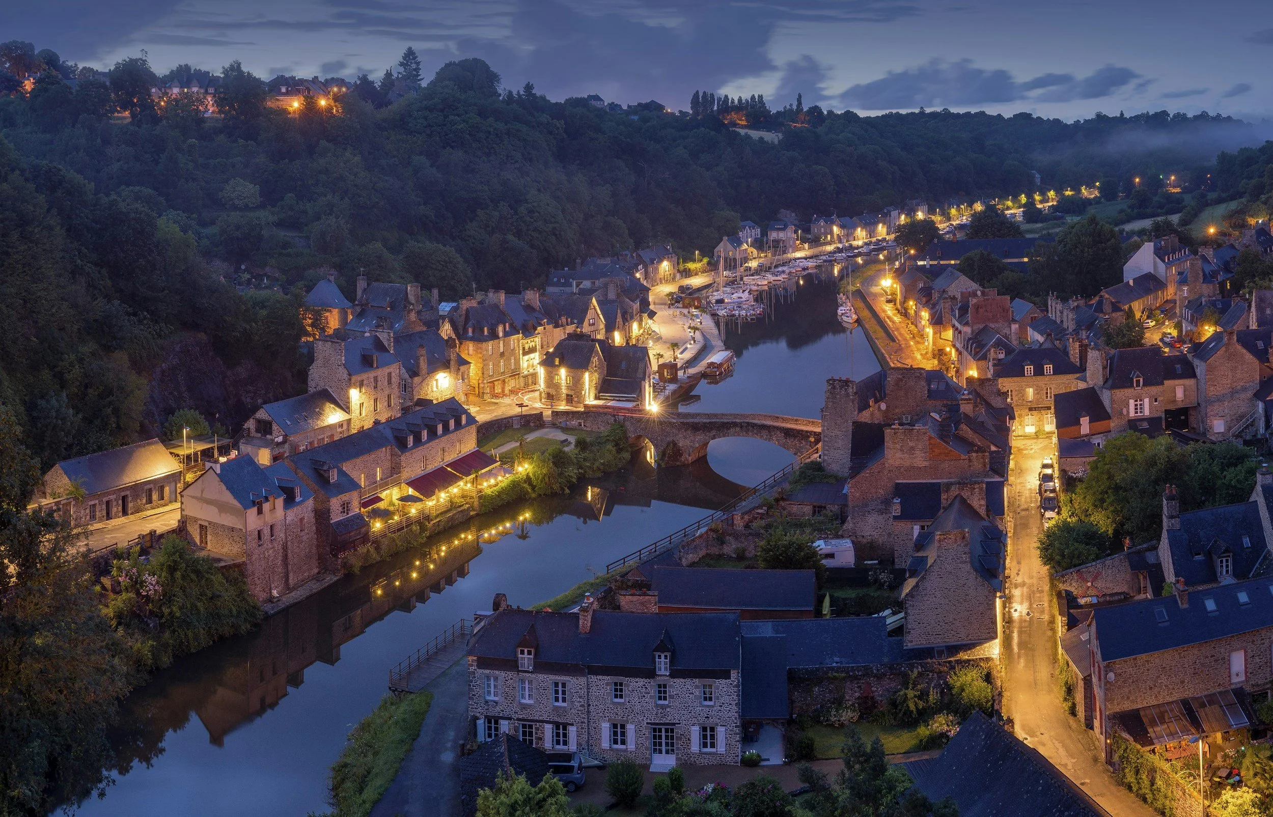 Aerial view of a small town at dusk with lit buildings, a river, and green hills in the background.