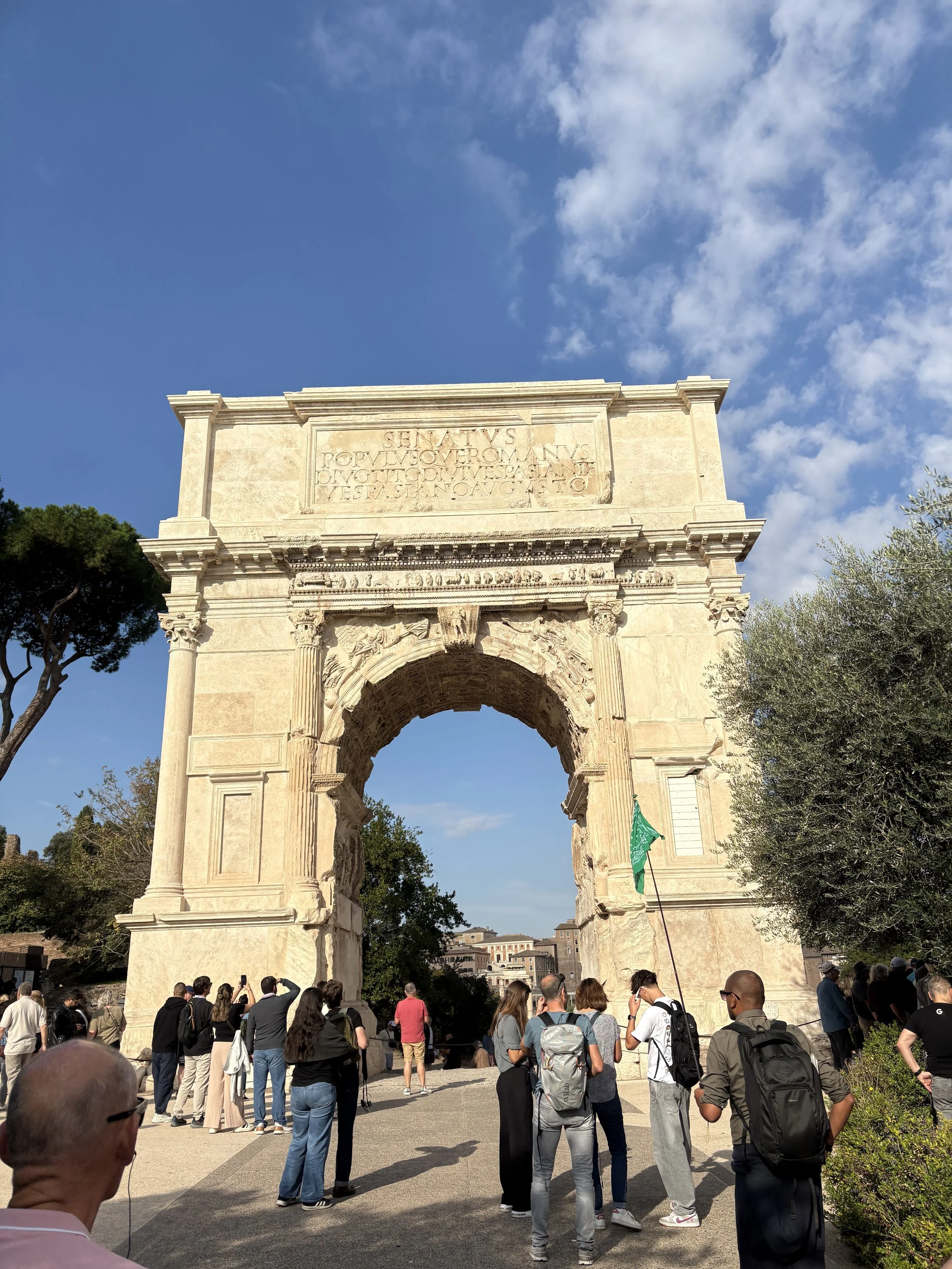 Ancient Roman triumphal arch with a crowd of tourists in front, blue sky with clouds, and trees on both sides.