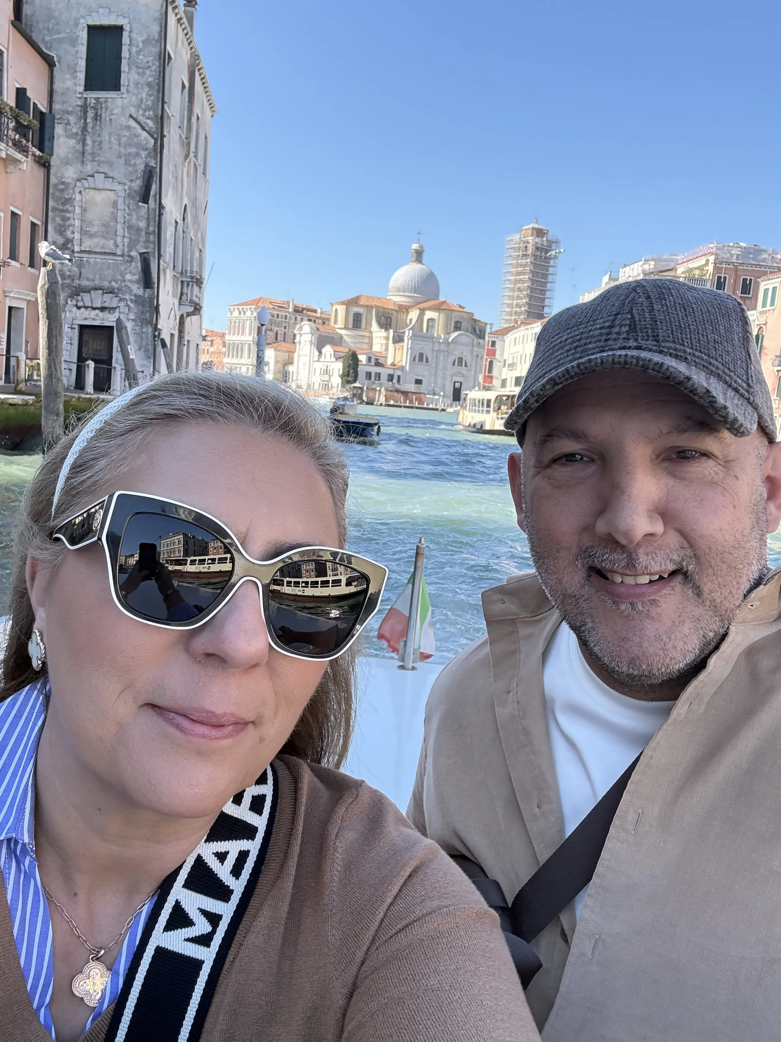 A selfie of a woman and a man on a boat cruising through a canal in Venice, Italy, with historic buildings and a church with a dome in the background.