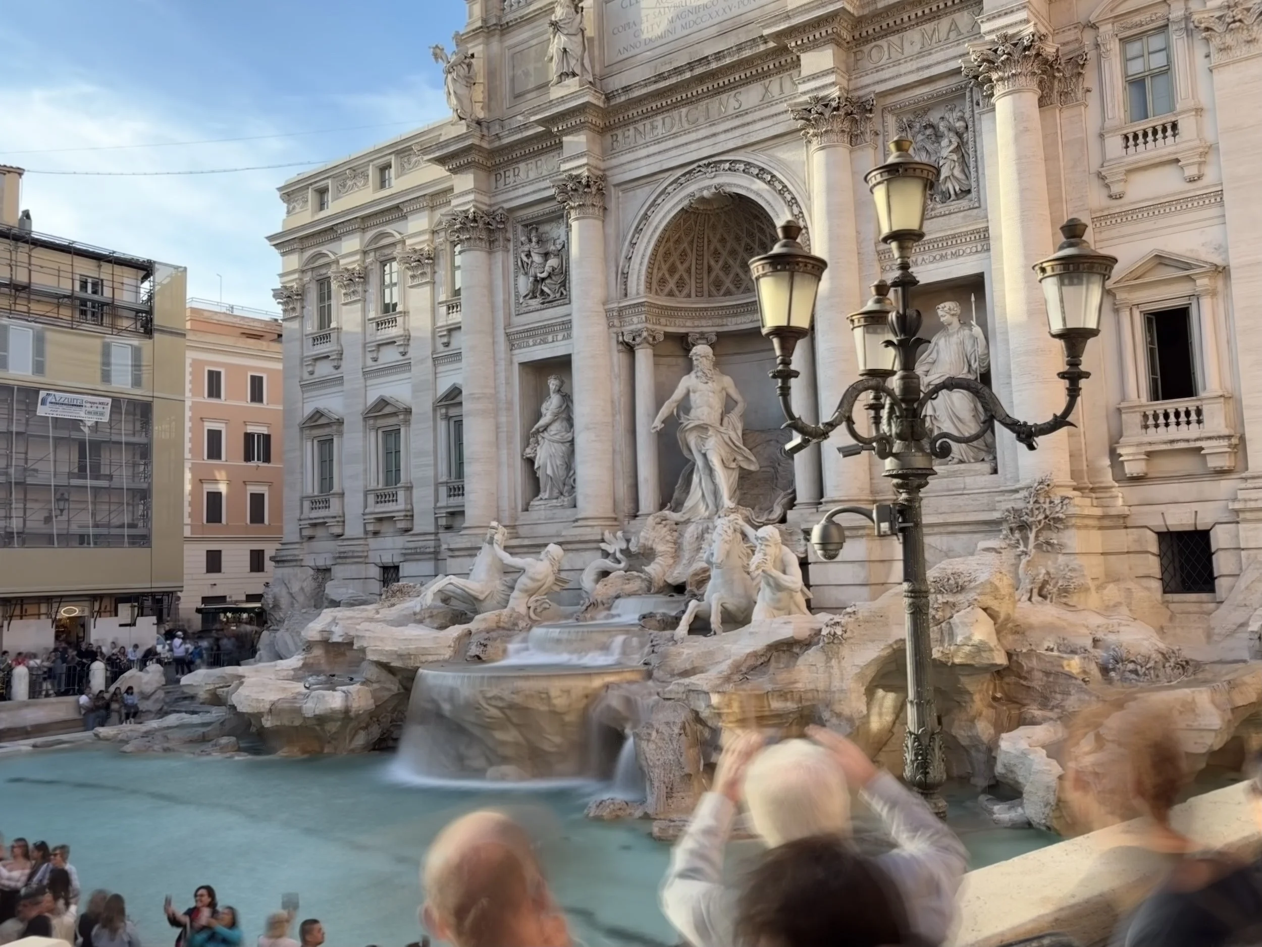 The Trevi Fountain in Rome, Italy, with ornate marble sculptures, statues, a cascading waterfall, and a large water basin, surrounded by buildings and a crowd of tourists taking photos.