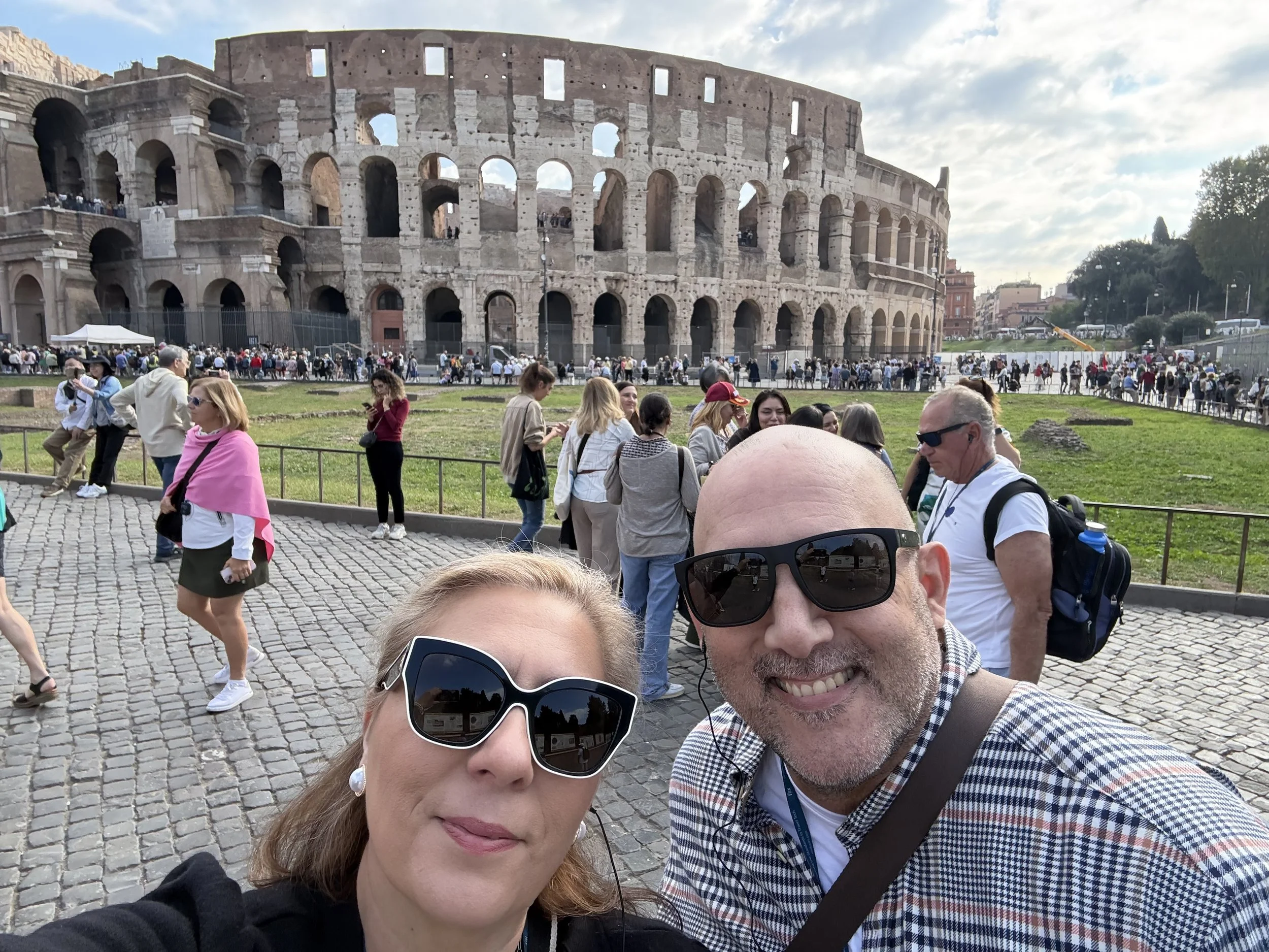 Two tourists taking a selfie in front of the Colosseum in Rome, Italy, with a crowd of visitors and the historic monument in the background.