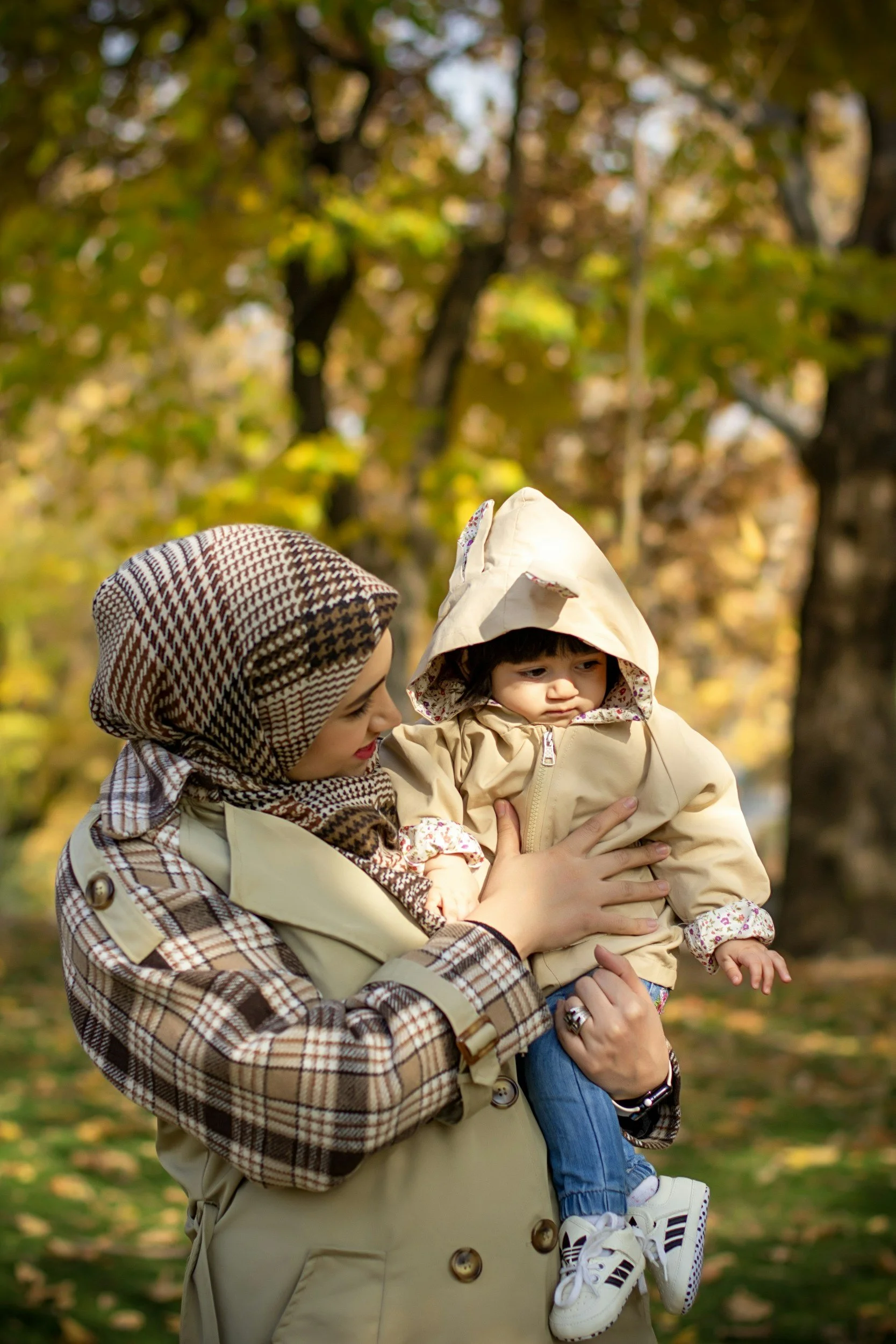 Woman holding a young child outdoors in a park during fall, surrounded by trees with yellow leaves.