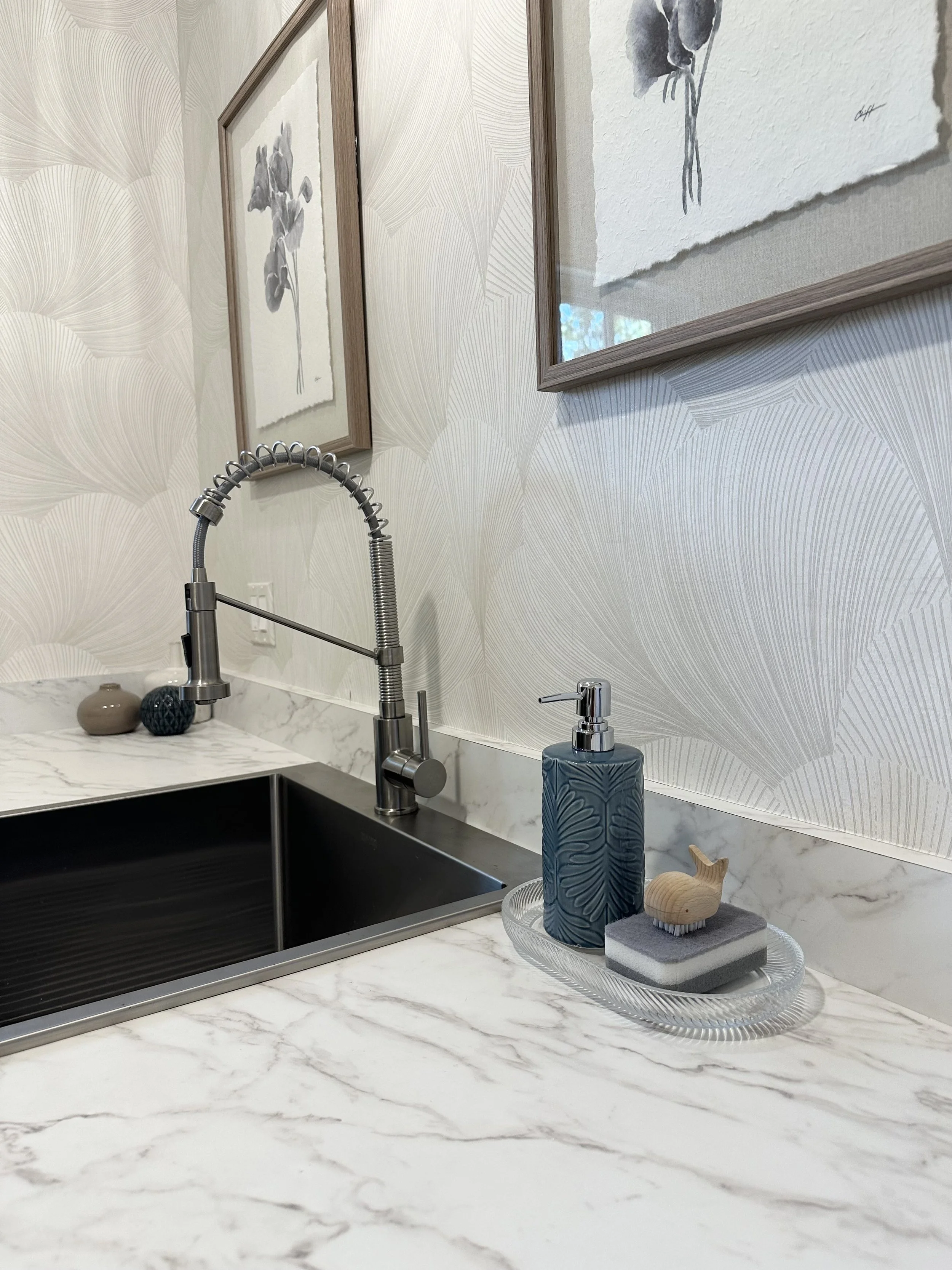 Bathroom countertop with a black sink, a silver faucet, a soap dispenser, a wooden whale-shaped figure, and a decorative tray with a nail brush. Two framed black-and-white botanical art prints hang on the patterned wallpaper behind.