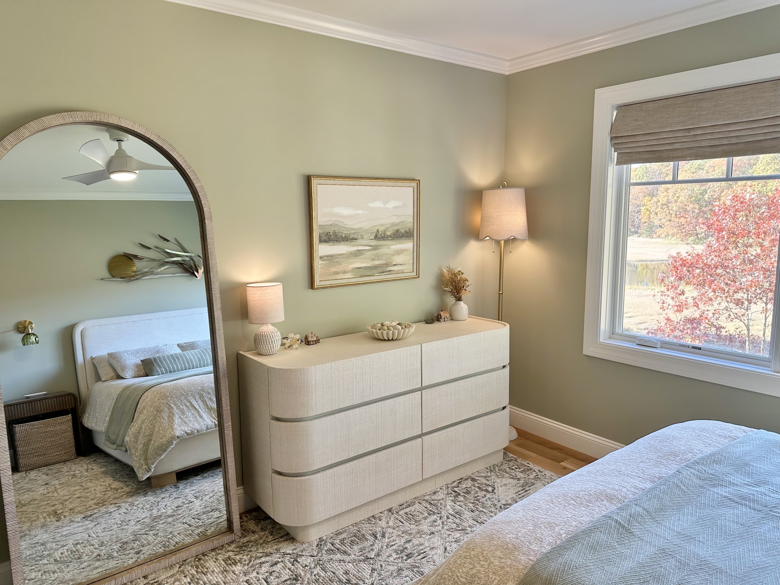 Bedroom with green walls, a dresser with decorative items, a window with a view of red trees, and a mirror reflecting a bed and ceiling fan.