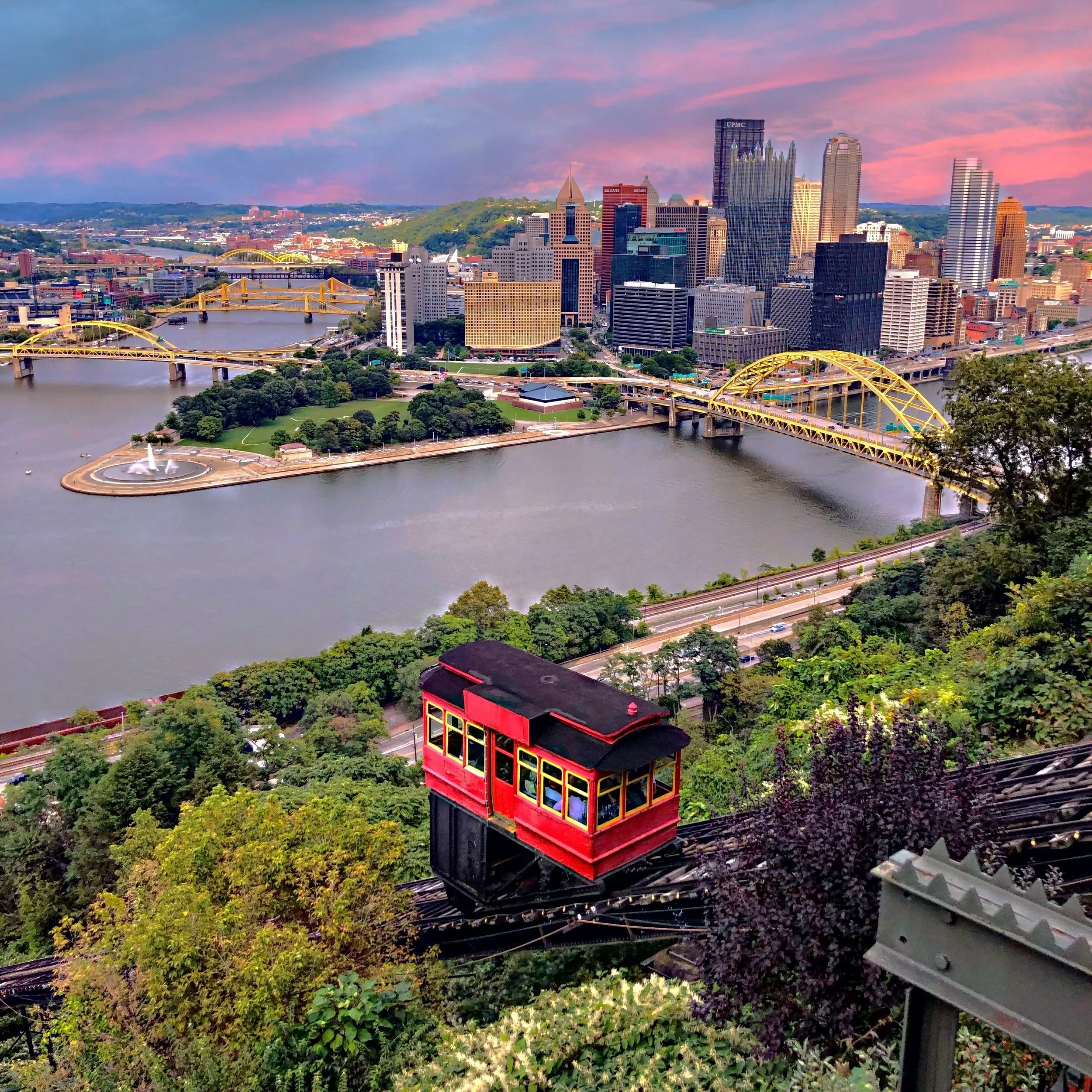 Pittsburgh city skyline at sunset with tall buildings, bridges over a river, a fountain on a small island, and lush green trees and the Duquesne incline trolley car in the foreground.